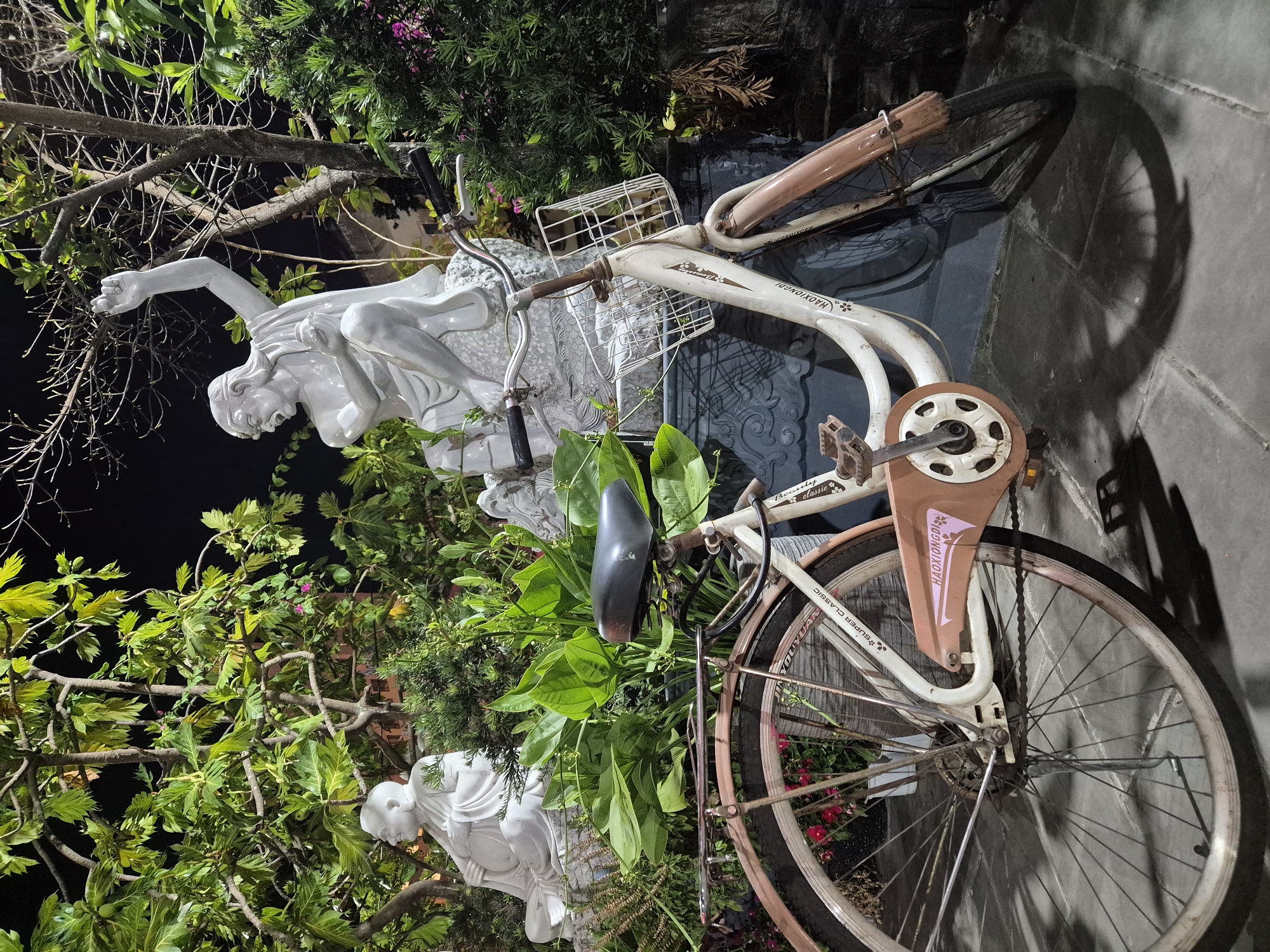 In Đà Nẵng, Vietnam, a white bicycle with a basket and a brown mudguard stands in front of a garden at night. Two statues are visible; one of a seated figure in white and another of a dynamic, gesturing figure also in white. Lush green foliage surrounds the statues and the bicycle, with branches of a tree reaching overhead. The ground is paved with stone tiles, and shadows are cast by the objects and plants, indicating artificial lighting. The bicycle shows signs of age with some rust on its components and is parked on the stone pavement. The scene appears to be a tranquil, perhaps spiritual, outdoor setting.