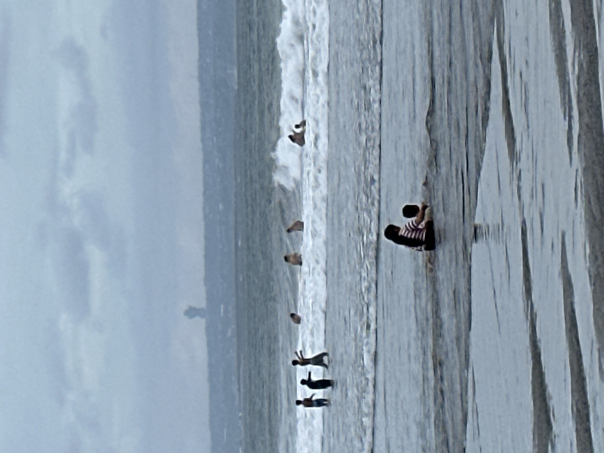 The image depicts a cloudy day at a beach in North Kuta, Indonesia. In the foreground, two figures, likely a woman and a child, are seated in shallow water. The woman is wearing a striped shirt, and the child is facing away from the camera. Further out in the ocean, several people are visible, some wading in the water and others playing in the waves. A distinctive structure resembling a monument or a tall building is faintly visible on the distant coastline. The water is choppy, with waves breaking near the shore and further out. The overall atmosphere is overcast and slightly hazy.