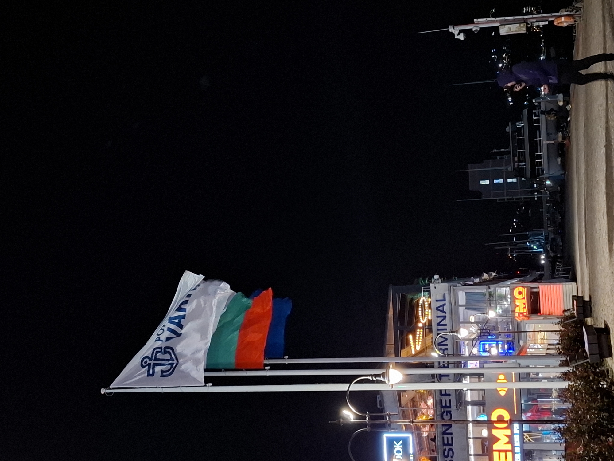 The image is taken at night in Varna, Bulgaria, near the harbor or port area. In the foreground, flags are visible, including one with a white background, a blue anchor, and the words "Port Varna." The other flags appear to be the Bulgarian flag (white, green, red) and potentially another flag (blue). These flags are mounted on poles.
Behind the flags, there is a building with neon signs and lights. Some signs are in Cyrillic script. The building appears to be a terminal or commercial establishment.
In the background, along a concrete walkway or pier, there are people walking. Beyond the walkway, there are structures that appear to be ships or parts of a marina, illuminated by lights. The sky is dark and clear.