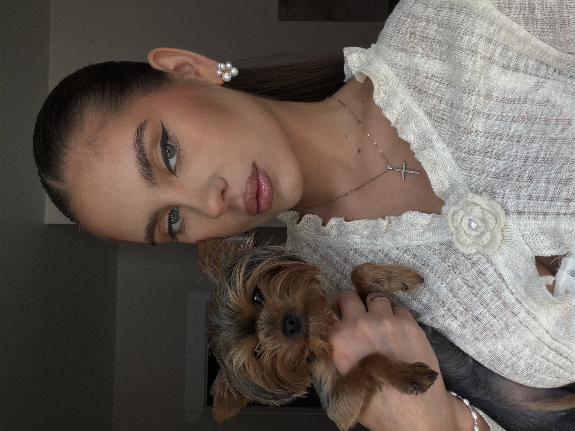 A light-skinned woman with brown hair pulled back tightly, wearing a white linen top with ruffle detailing and a pearl flower embellishment, holds a Yorkshire Terrier dog. The woman has visible winged eyeliner and is wearing pearl earrings and a delicate gold necklace with a cross pendant. Her expression is neutral as she looks toward the camera. The dog is nestled in her arm, looking forward with brown eyes. A silver bracelet is visible on her wrist, and a ring is on her finger. The background is blurred, but suggests an indoor setting with neutral tones, including a wall painted in a dark color on the left side of the image. There is no specific evidence to place the image in Krasnoyarsk, Russia.