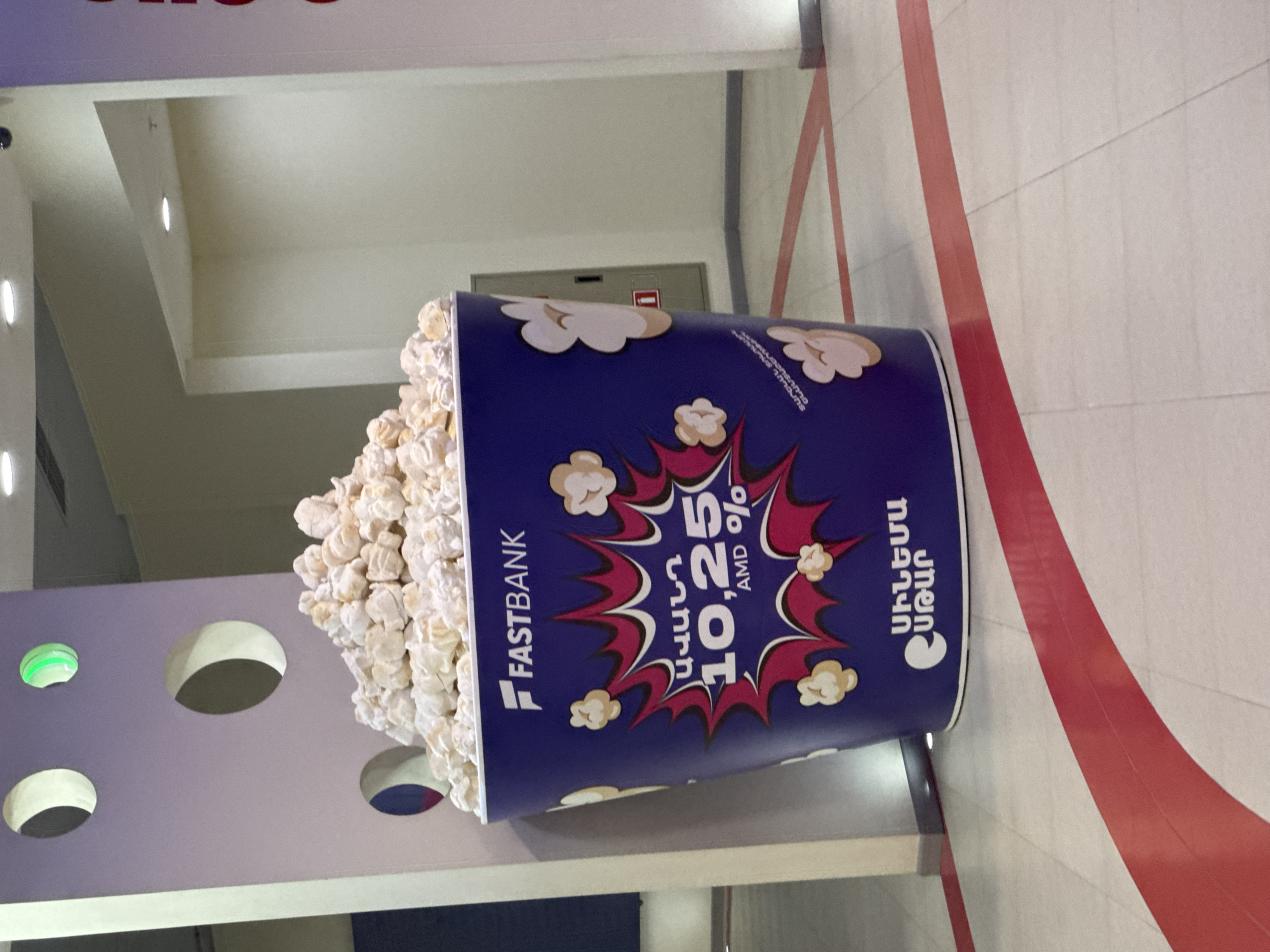 This image displays an oversized, cylindrical popcorn bucket serving as an advertising display within an indoor, well-lit commercial space, likely in Yerevan, Armenia. The bucket, oriented upright, is predominantly dark blue with a white top edge, and is filled with a large amount of white, textured popcorn.
The bucket's design features the "FASTBANK" logo vertically on its left side. A prominent, comic book-style explosion graphic in red and white occupies the central part of the bucket, containing the numbers "10,25 AMD %" and Armenian text. The Armenian text includes "ԱՆՆԱ ԱՄԱՆ" (Anna Aman) above the percentage, and "ՍՊԻՆ ՍՊՈՐՏ" (Spin Sport) below it, suggesting an offer or campaign name from FastBank. Illustrations of cartoon popcorn kernels are scattered across the blue surface of the bucket.
The surrounding environment includes a light-colored tiled floor with wide, curved red stripes that define a path or design element. The walls are also light-colored, with a section to the left featuring multiple circular cutouts, one of which emits a green light. The ceiling is white with visible recessed lighting. The scene is static, devoid of people, and appears to be in a modern public building such as a mall, cinema lobby, or bank branch.