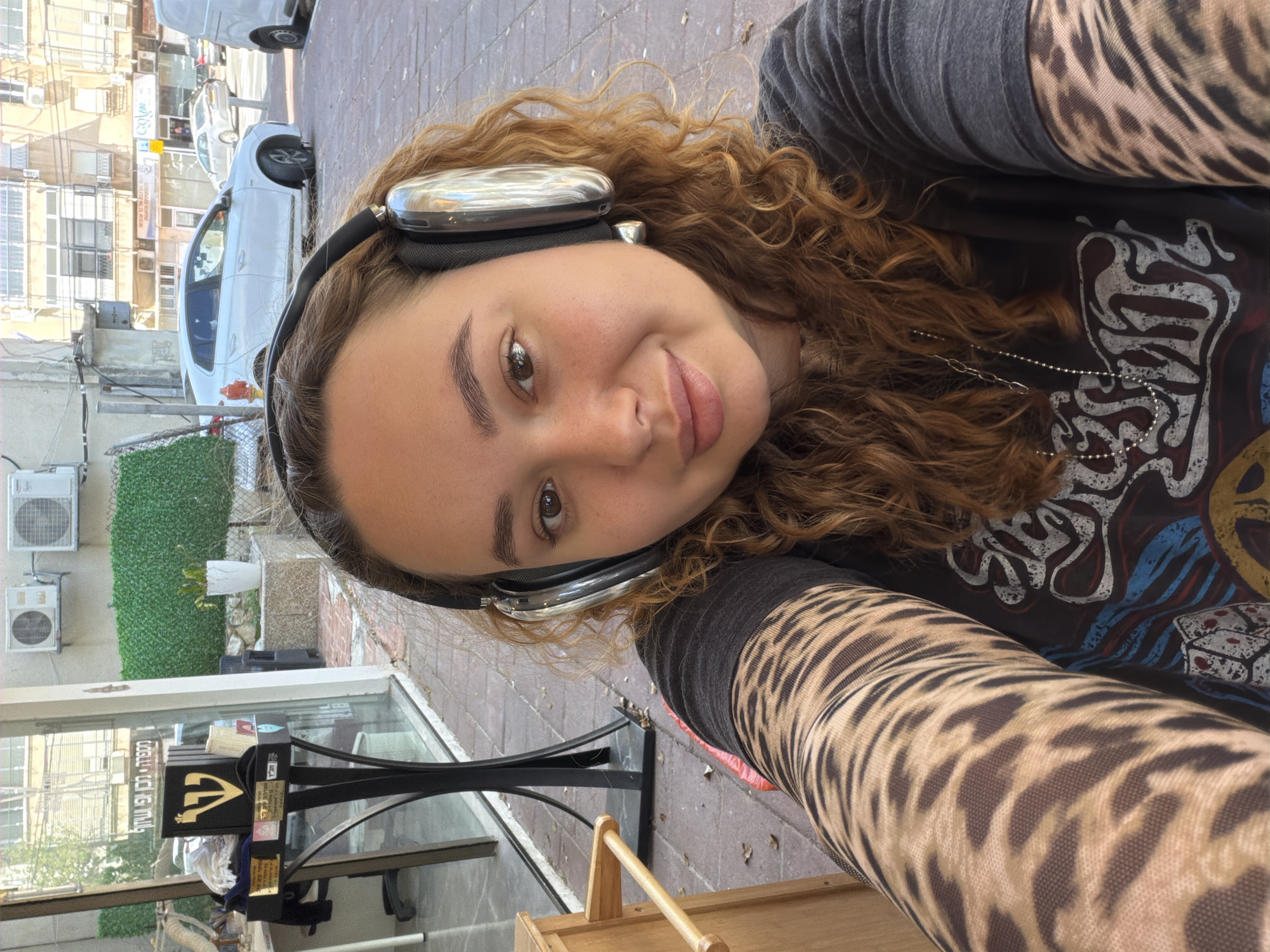 A selfie taken in Ramat Gan, Israel, shows a young woman with curly auburn hair wearing silver headphones. She is wearing a dark t-shirt and tiger-striped patterned sleeves. In the background, there is a street scene with cars, buildings, and potted greenery. A black signboard with Hebrew writing is visible in the foreground next to a window.