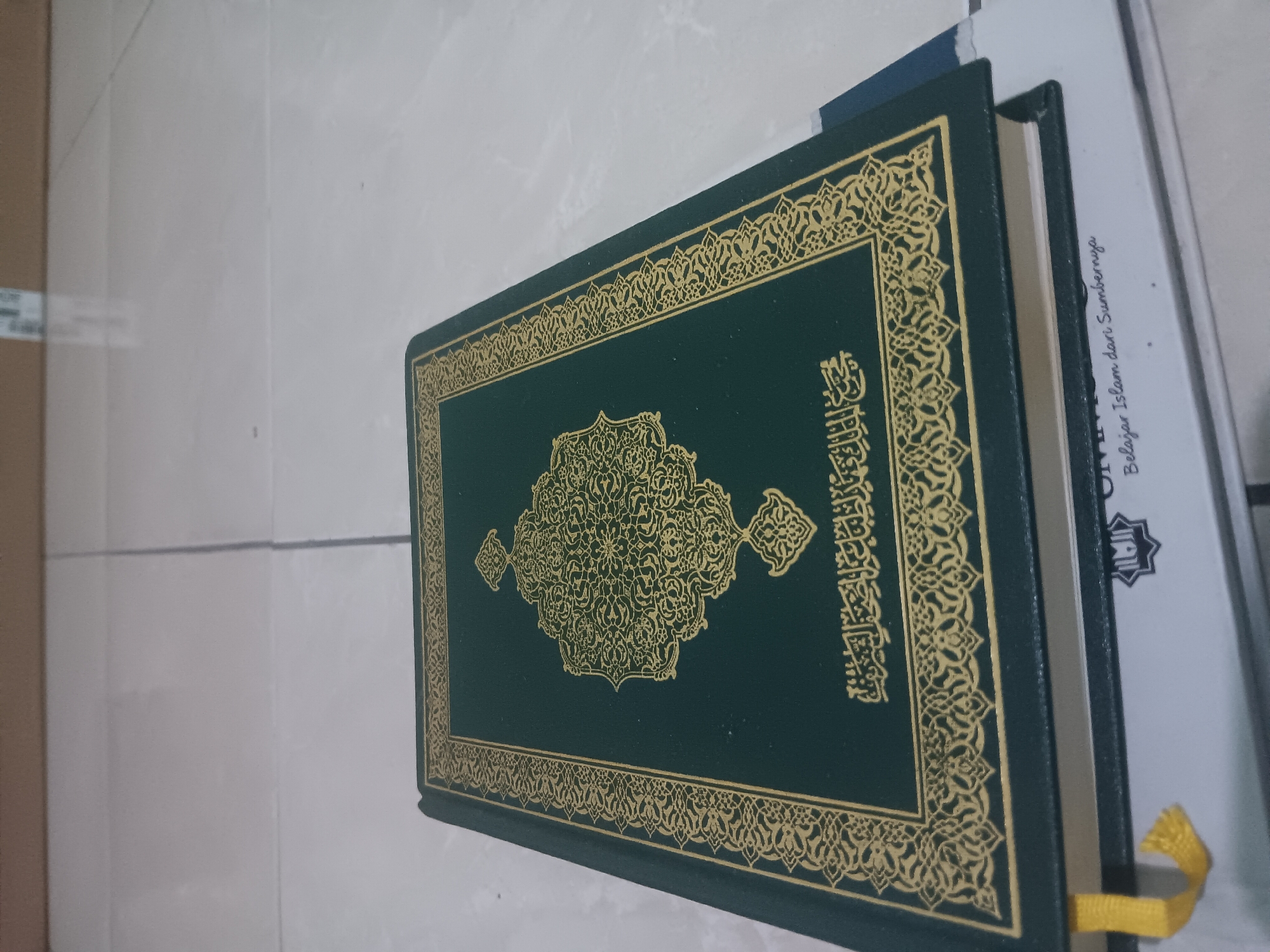 A closed, dark green book with gold Arabic calligraphy and intricate designs on its cover rests on a light-colored tiled surface. A yellow ribbon bookmark hangs from the left edge of the book. Beneath the dark green book, a lighter-colored book with text and a star-like symbol is partially visible. The tiled surface appears to be indoors, likely a floor. The image does not contain enough information to determine the specific location within Batursari, Indonesia.