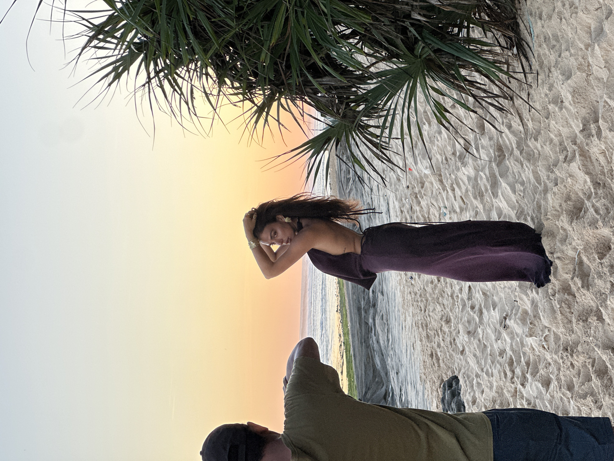 A vertical photograph captures a scene at a beach in Kuta Selatan, Indonesia, during sunset. A woman with long, dark hair stands in the middle of the frame; she's wearing a dark, draped garment. She poses with her hands near her hair. Below her and to the front, a man wearing an olive-green shirt and a dark baseball cap is partially visible, implying he is the photographer. The beach sand covers the ground, and the waves gently lap at the shore in the background. Above the woman, the fronds of a beach palm tree create a natural frame, casting a shadow over part of the scene. The sky transitions from light to orange at the horizon.