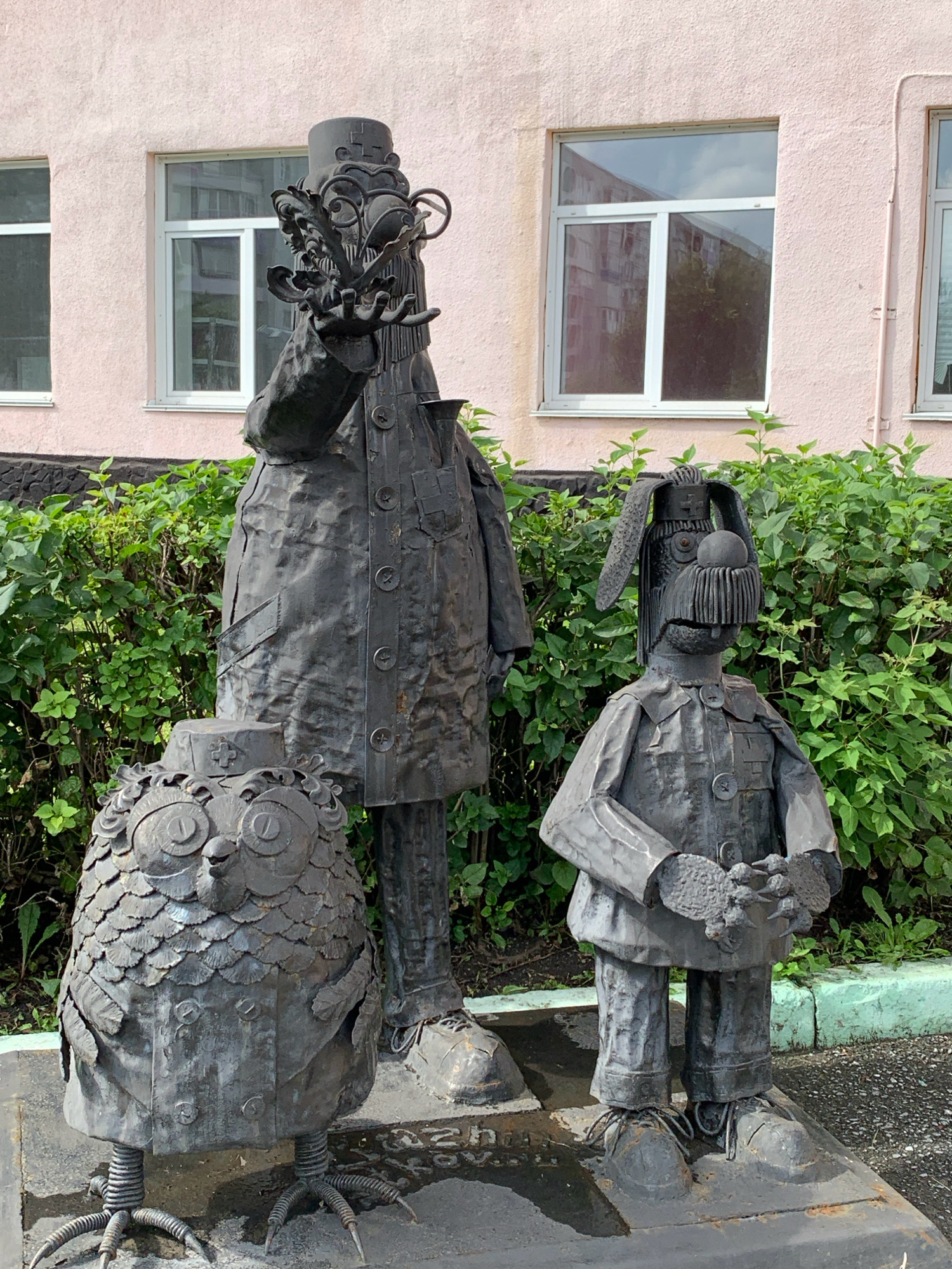 The image depicts a collection of three metal sculptures in front of a pale pink building with white-framed windows. The sculpture on the left is an owl with large, circular eyes, a hat resembling a small cap with a cross, and feathered detailing on its body. It stands on thin, wire-like legs with clawed feet. To its right is a tall, slender figure wearing a hat with a rounded top and a decorative ring, a long coat with multiple buttons, and an ornate facial structure with a large nose and beard-like protrusions. This figure has one hand extended, holding a cluster of leafy objects. The third sculpture, to the right of the tall figure, is a smaller, rabbit-like character with long ears, a round nose, and a multi-buttoned jacket. It is holding a small, textured object in its hands. All three figures appear to be made of weathered, dark metal. Behind the sculptures is a dense hedge of green bushes, and the ground in front of them is paved. The scene is located in Prokopyevsk, Russia, at the entrance of the Prokopyevsk State Technical College.