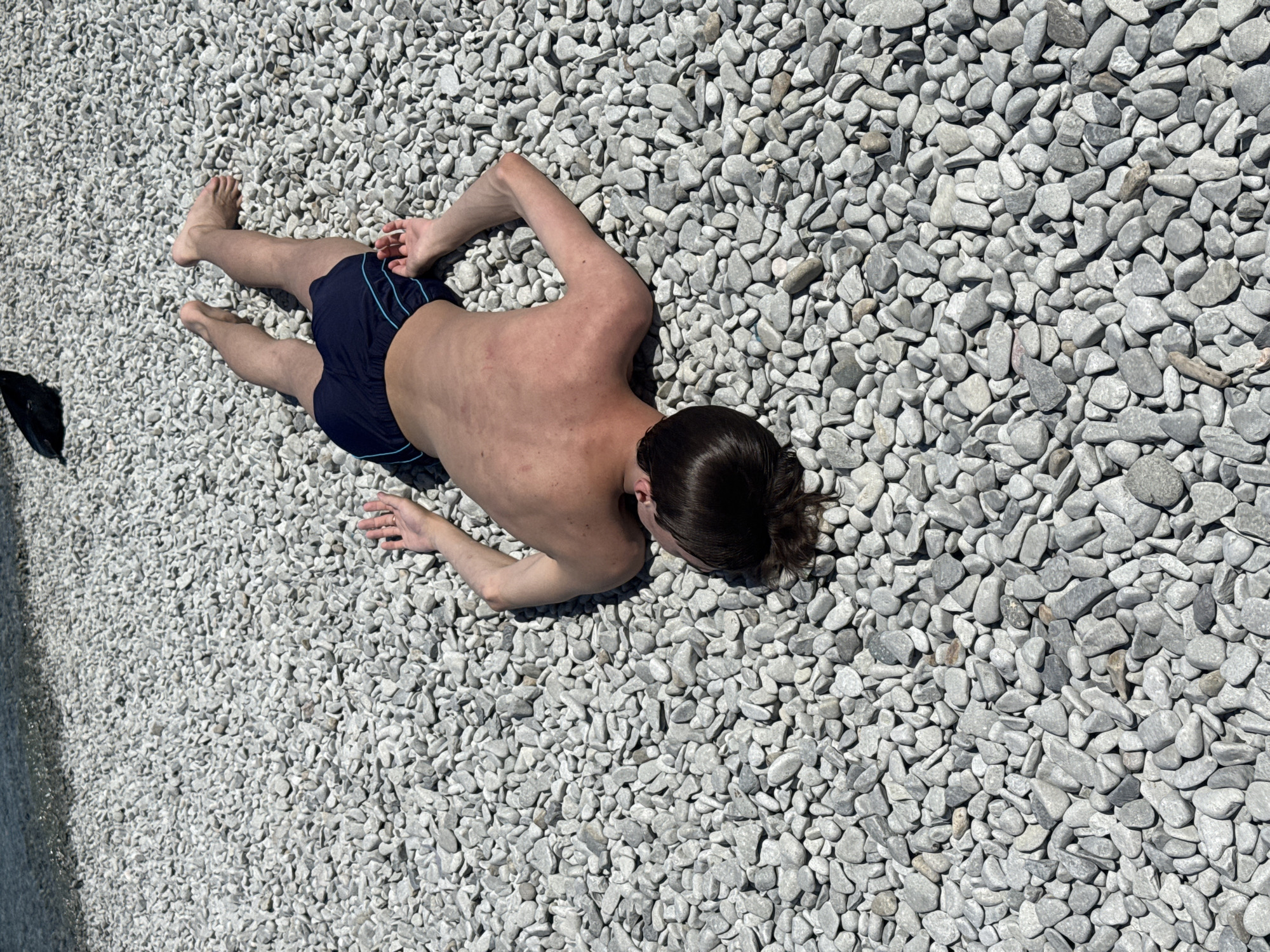 A person is lying on their stomach on a beach covered in small, gray pebbles. The person is wearing dark blue swim trunks with a light blue trim. Their back is facing the camera, and they appear to be sunbathing. The sun is bright, casting shadows on the pebbles, suggesting it is daytime. The scene is set on a pebbly beach, possibly near a body of water, though the water itself is not clearly visible. The overall mood seems relaxed and leisurely, typical of a day at the beach. There is no visible text in the image.