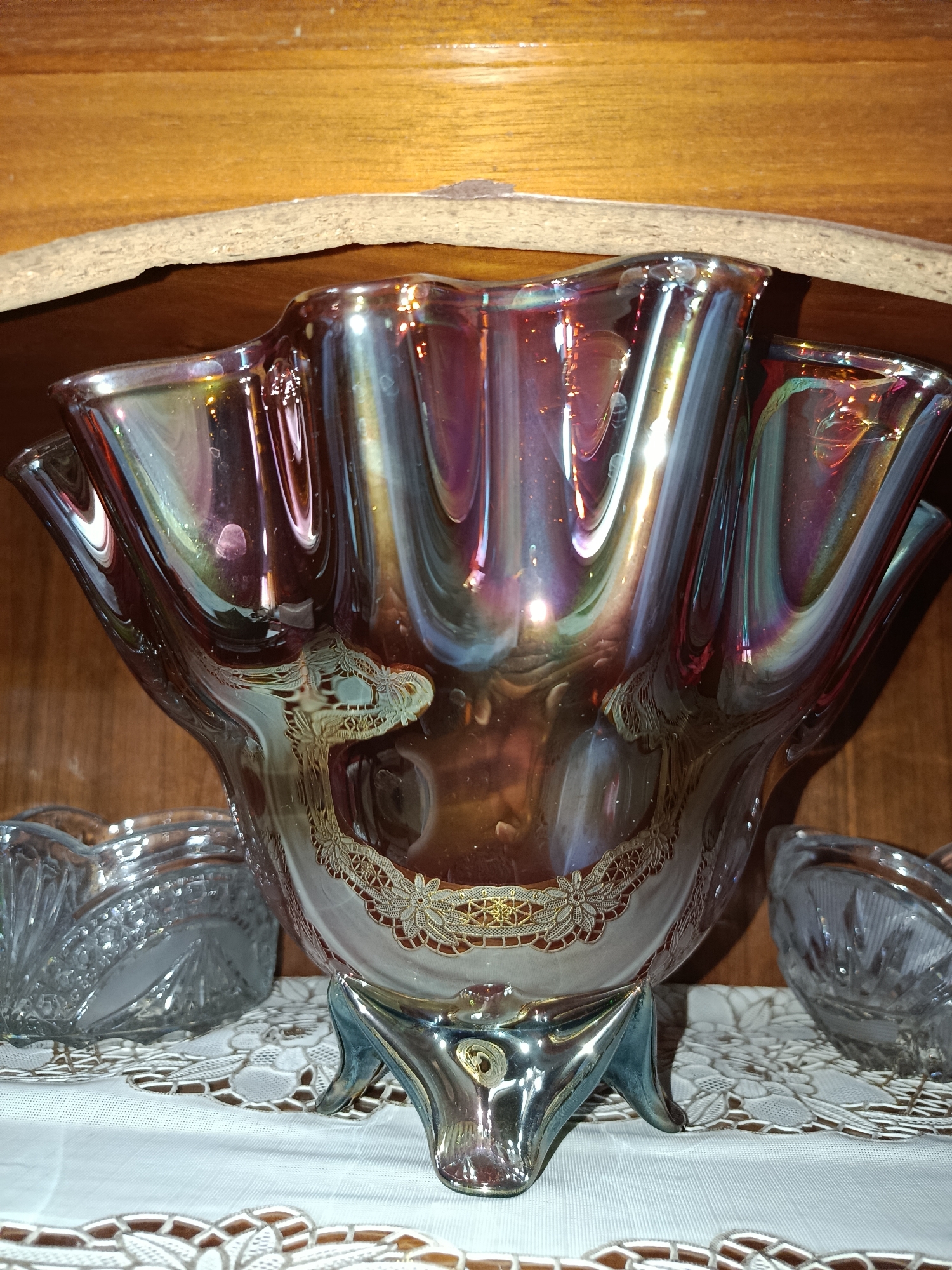 The image shows a close-up of an iridescent glass bowl with ornate detailing, resting on a white lace doily. The bowl has a wavy rim and sits on three decorative feet. To the left and right of the main bowl are portions of two smaller, clear glass containers with faceted designs. The objects are displayed on a white fabric surface, likely a tablecloth or shelf covering. Behind the display is a wooden surface, possibly a shelf or cabinet interior, with horizontal planks. The lighting creates a shimmering effect on the glass. The scene is indoors, set within a domestic or display environment, but there is no specific indication of its location within Tashkent, Uzbekistan.