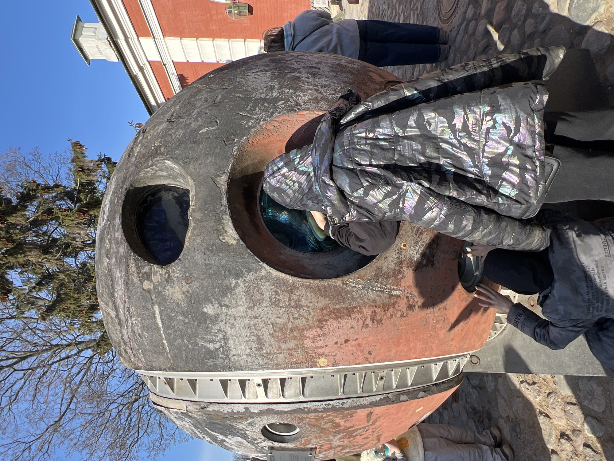 The image shows a spherical, weathered metallic object with circular openings. People are interacting with the object, looking inside and touching it. The object appears to be a spacecraft capsule of some kind, with visible wear and tear. The environment includes bare trees, a red and white building in the background, and a cobblestone ground surface. The location is in the city of Saint Petersburg, Russia.