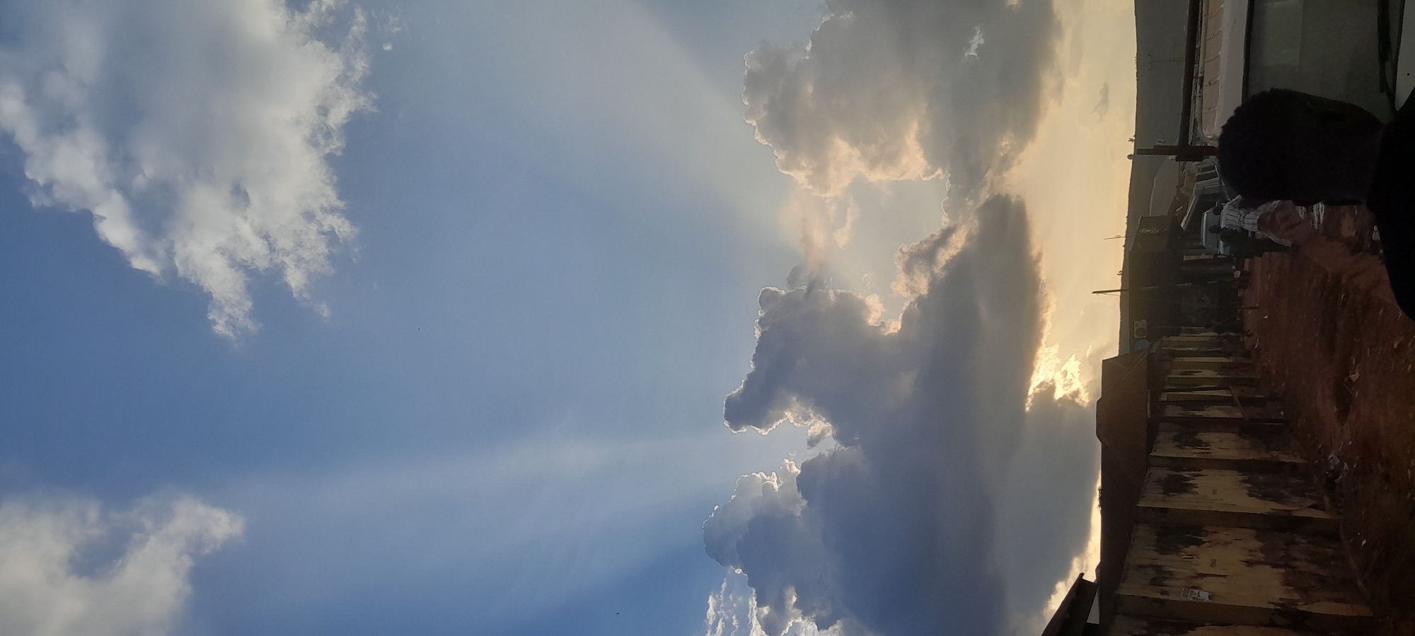 This photograph captures a dramatic sky over a street scene in Abuja, Nigeria. The sky is dominated by a mix of dark, voluminous clouds and bright, sunlit clouds. Rays of sunlight are visible, piercing through gaps in the clouds, creating a crepuscular effect. In the foreground, a rough, dirt road leads past a row of weathered, single-story buildings with corrugated metal roofs. The walls of these buildings are textured and appear aged. Beyond the buildings, vehicles and several individuals are present on the street, some of whom appear to be interacting or moving about. The overall impression is one of a busy, informal urban environment under a striking sky. The presence of specific architectural styles and the general atmosphere suggest a typical street in a developing area of Abuja, likely a marketplace or residential neighborhood.