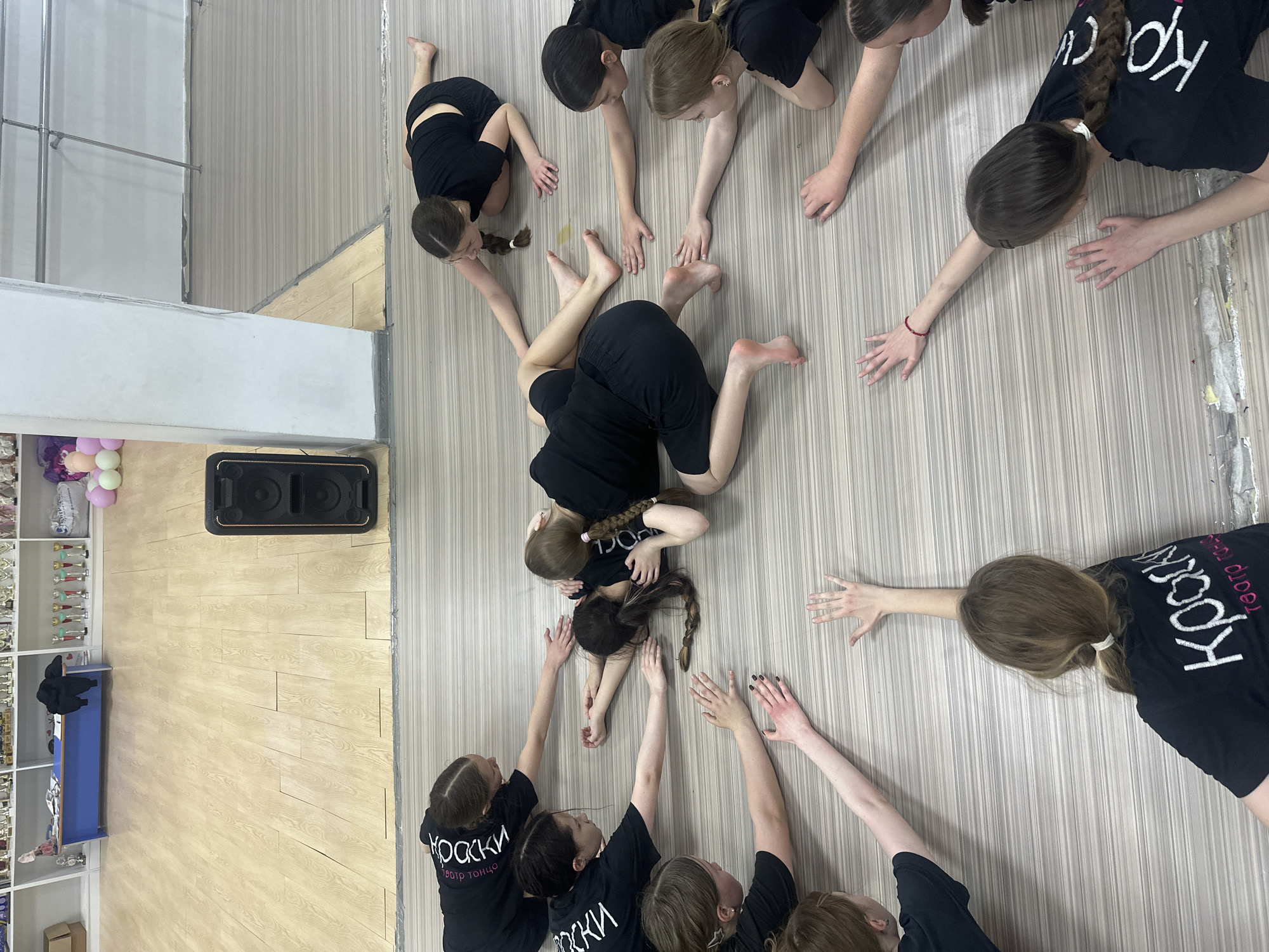 A group of young women, likely dancers, dressed in black shirts with white lettering, are arranged in a circle on a light-colored wooden floor inside a room. The word "Kраски" (Paints) and "Театр танца" (Dance Theater) are visible on some of their shirts. The women are reaching their arms towards the center of the circle. Two women are lying in the center, intertwined. A ballet barre is visible on the left side of the image, along with a wall shelf holding trophies. A sound speaker is also visible. The location appears to be a dance studio in Votkinsk, Russia.