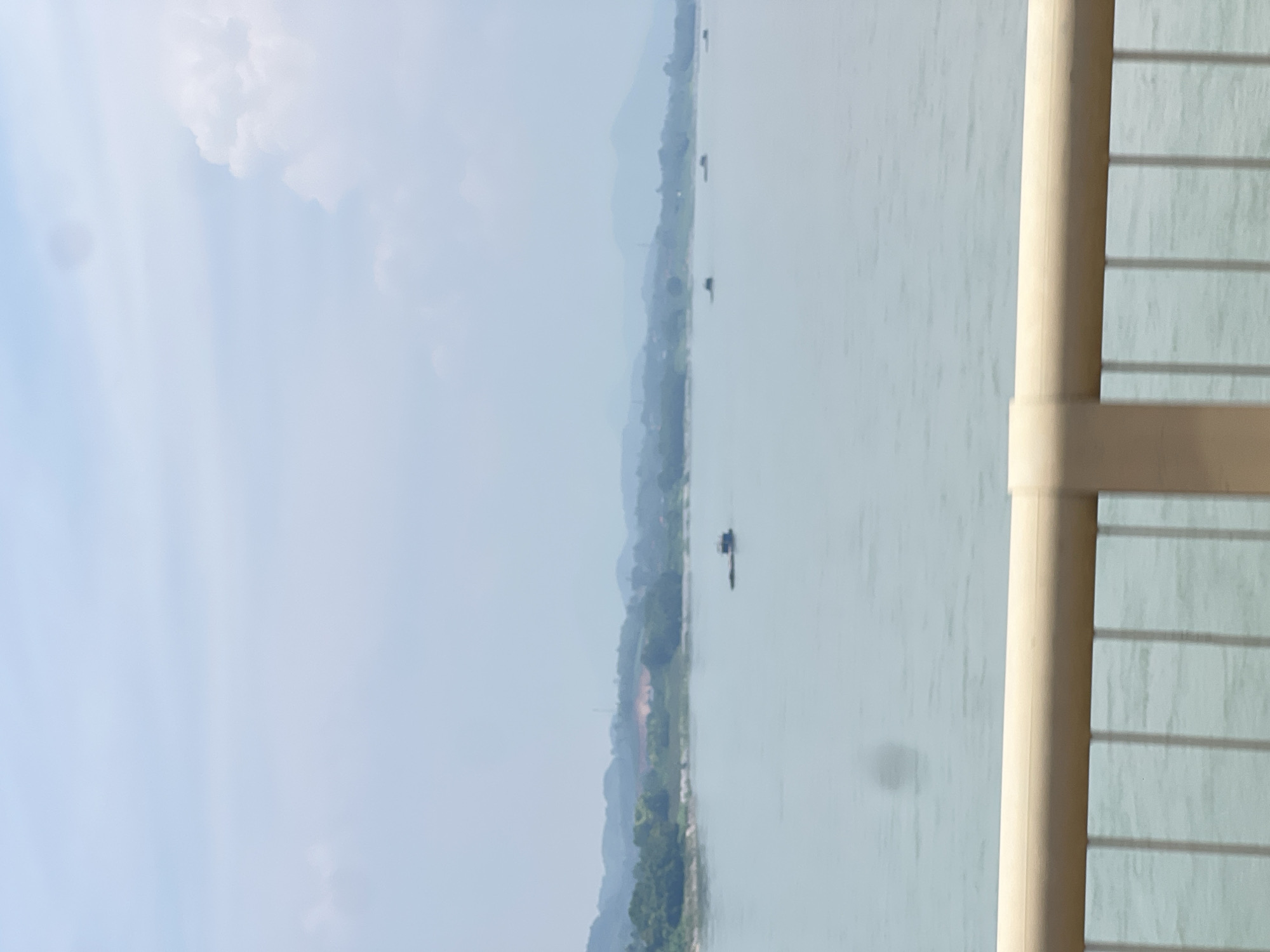 A wide, hazy shot captures a serene river scene in Hà Nội, Vietnam. The foreground features a beige railing and vertical metal bars, suggesting the viewpoint is from a bridge or elevated walkway. Beyond this, the calm, light-blue water of the river stretches out, with several small, dark boats scattered across its surface. The middle ground shows a distant, green, tree-lined shore, with rolling hills and mountains fading into the misty blue horizon. The sky is a pale blue, with soft white clouds, creating an overall atmosphere of tranquility and expanse. There are no people visible in the image.
