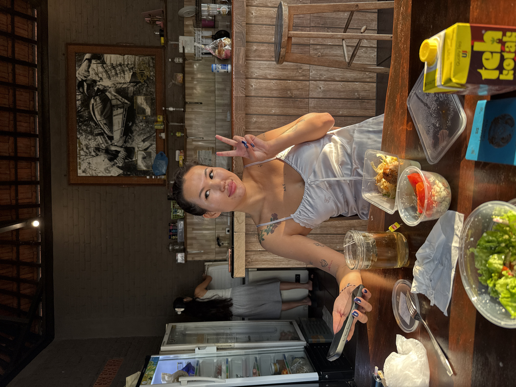 A young woman with dark hair and tattoos is seated at a wooden table in an open-air establishment with a thatched roof. She is wearing a light-colored slip dress and holding a smartphone in her left hand while making a peace sign with her right. In front of her are several takeout containers with food, a glass of iced tea, and a fork and napkin. Behind her, a woman in a white tank top and grey shorts is standing by a refrigerator. The background shows a brick wall with a large framed picture and shelves stocked with various items. A wooden stool is visible to the right of the woman. The establishment appears to be located in North Kuta, Indonesia, given the presence of "Teh Kotak," a popular Indonesian iced tea brand, on the table. The lighting suggests it is late afternoon or early evening.