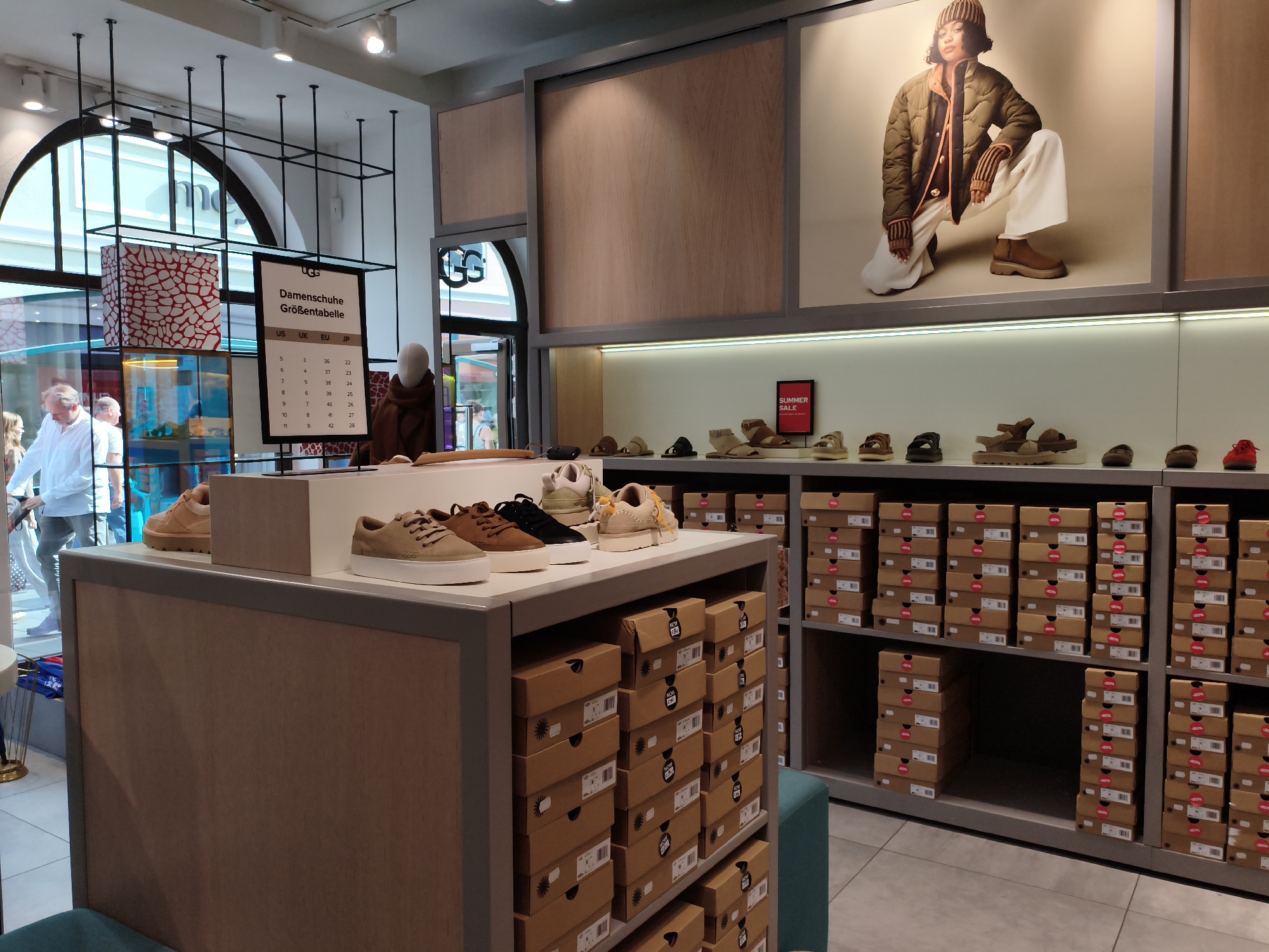 The image shows the interior of a UGG shoe store in Neumünster, Germany. The store features a well-lit display area with shoes and shoe boxes. In the foreground, a display case showcases several pairs of UGG shoes, including sneakers and sandals, in various colors such as beige, brown, and black. Behind the shoes, there are stacks of brown UGG shoe boxes, many of which have red sale stickers indicating a discount.
To the left, a large poster on the wall displays a young woman wearing a green puffer jacket and white pants, posing in a squatting position. Below the poster, a shelf displays more shoes and a "SUMMER SALE" sign. Further to the left, there is a glass display with colorful patterned boxes and a size chart for women's shoes.
In the background, through a glass doorway, other customers and employees are visible, suggesting a busy retail environment. The overall impression is of a modern and organized shoe store.