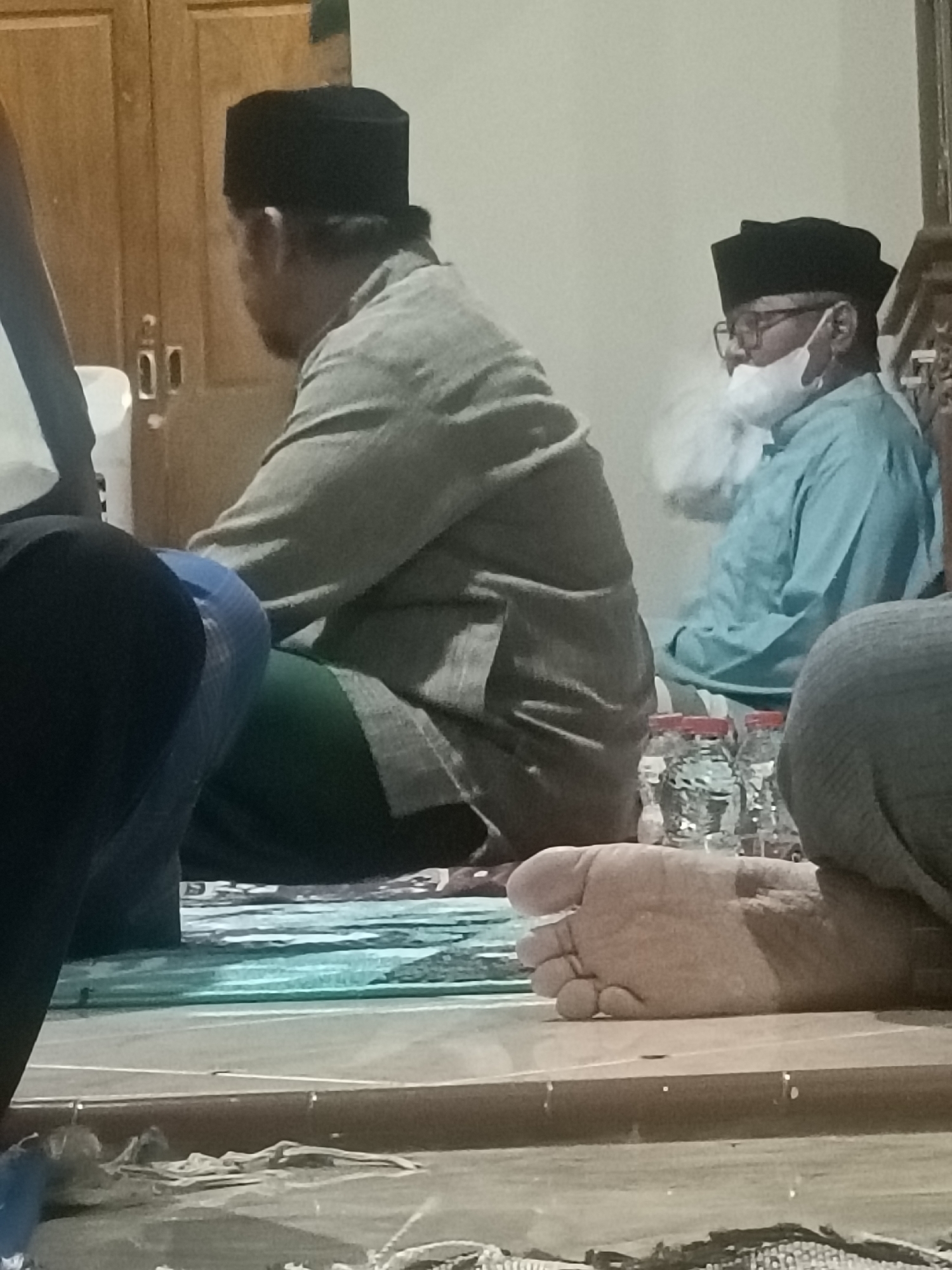 The image shows three people sitting indoors, presumably in Kediri City, Indonesia. In the foreground, a person's bare foot is visible, resting on a flat surface. Behind this foot, two individuals are seated, facing away from the camera. The person on the left wears a gray long-sleeved shirt and a black skullcap. The person on the right wears a light blue long-sleeved shirt, glasses, and a white mask. Behind them, a wooden door with two vertical panels is visible. In the mid-ground, three plastic bottles with red caps are placed on a surface. The environment appears to be a room with light-colored walls and a wooden floor.