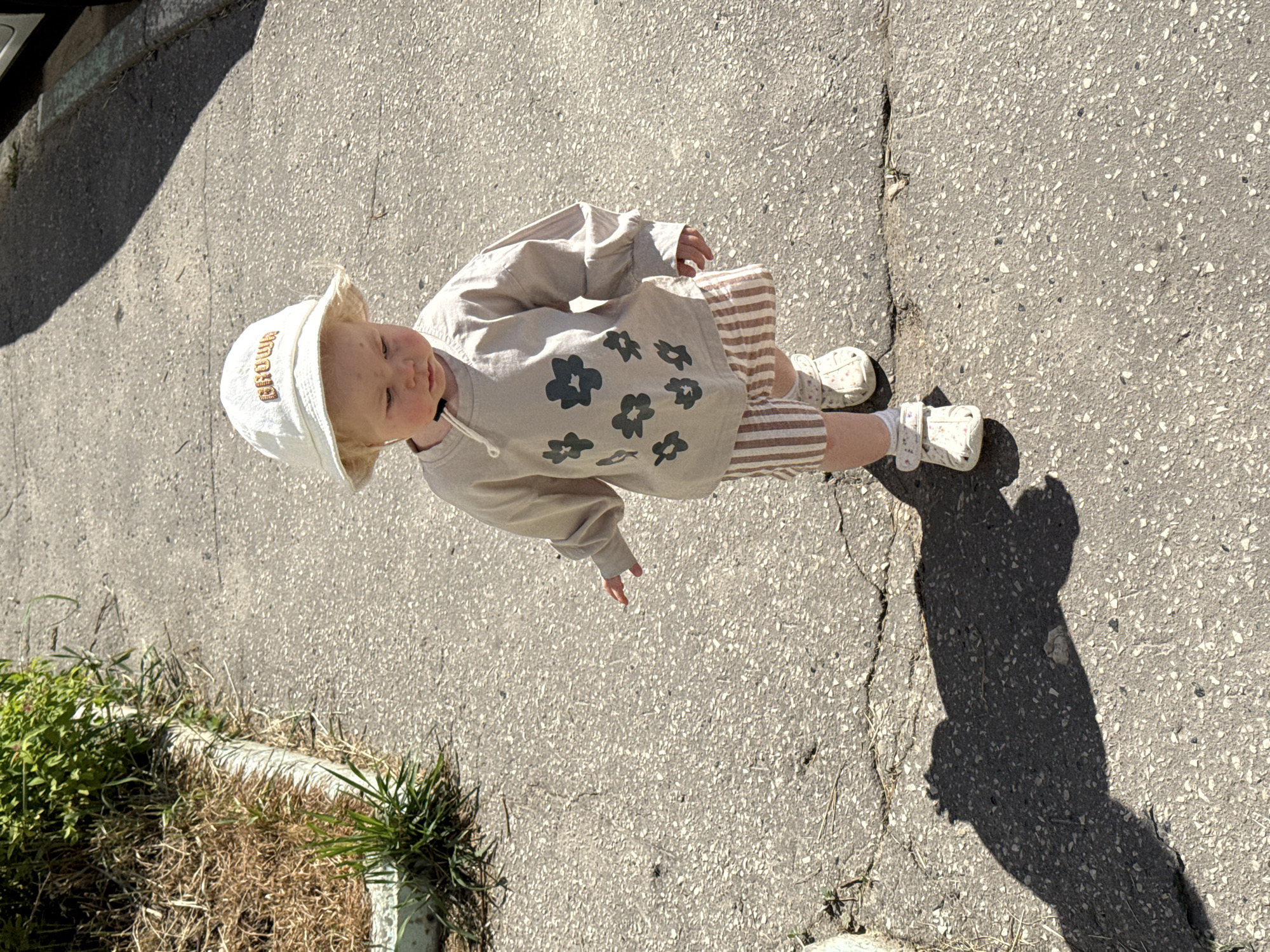 A high-angle, full shot depicts a toddler standing on an asphalt surface. The toddler wears a light-colored hat with text embroidered on the brim, a long-sleeved beige shirt with a floral design, and striped beige and white shorts. Small white shoes are visible on the toddler’s feet. The asphalt surface is cracked in places. Vegetation grows near the edge of the asphalt. The toddler's shadow is cast on the asphalt. The location is not specified in the prompt. Therefore, the location in the city of Vladimir, Russia, cannot be confirmed.