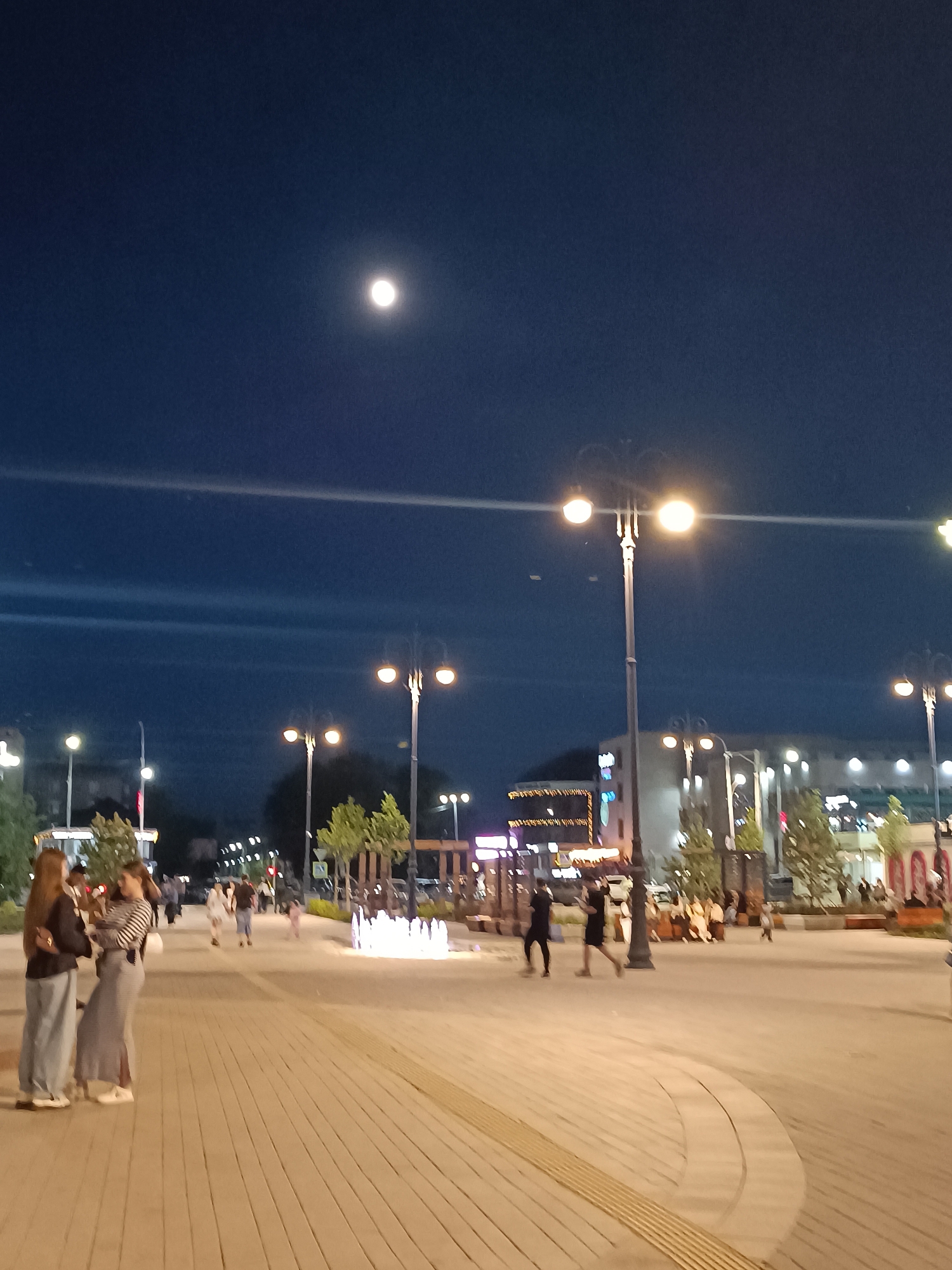 At night in Kokshetau, Kazakhstan, a brightly lit city square features numerous individuals strolling along a brick-paved pathway. In the foreground, two women converse. In the background, a row of streetlights illuminates the scene. A full moon is visible in the night sky.