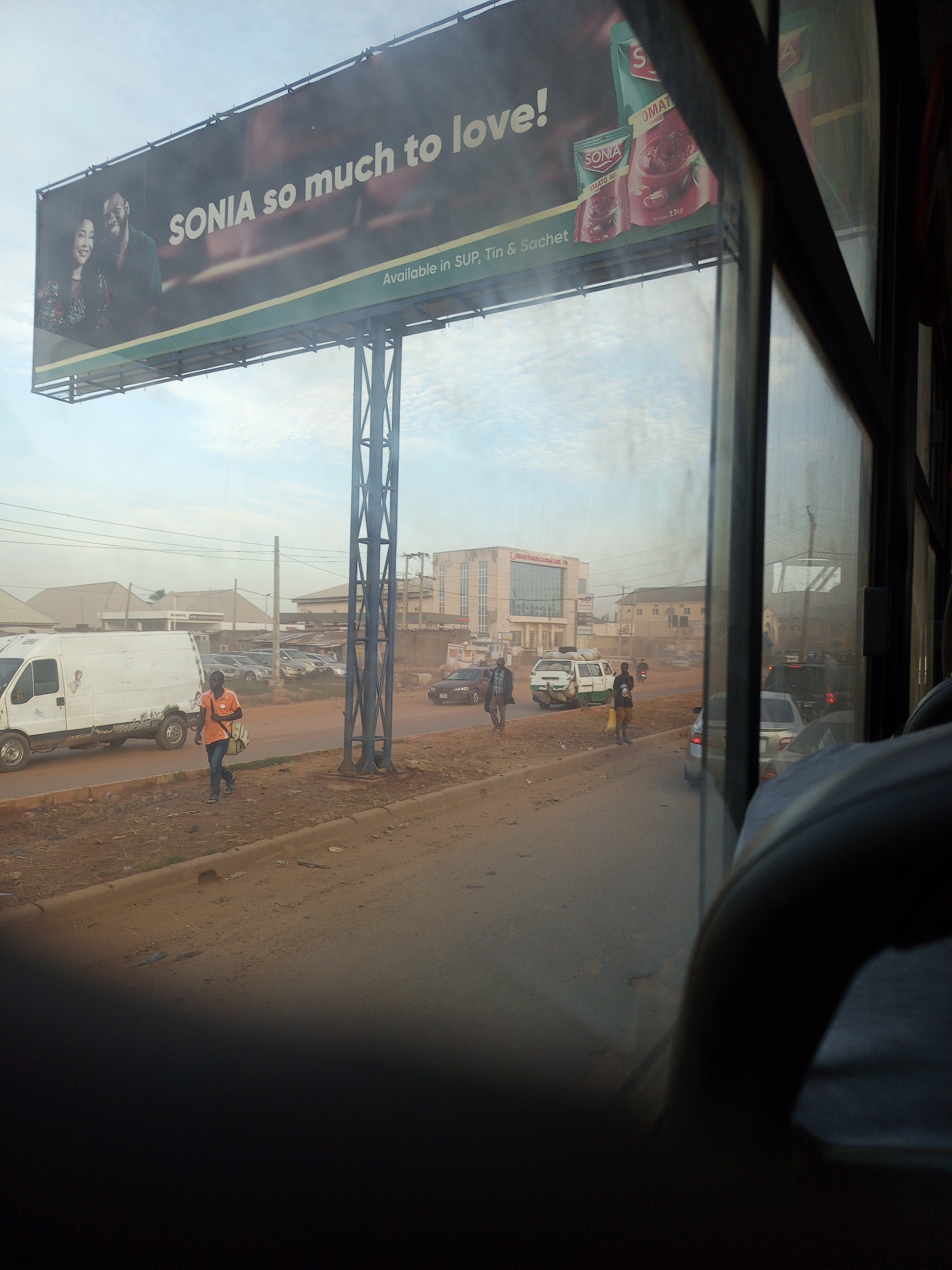 The image depicts a view from inside a vehicle, likely a bus, looking out at a street scene in Ado, Nigeria. A large billboard for "SONIA" with the slogan "so much to love!" is prominent in the upper portion of the image. The billboard features an image of a couple and product packaging for "SONIA TOMATO M." In the foreground, a man in an orange shirt walks along the side of the road carrying a light-colored bag. Several vehicles, including a white van and other cars, are present on the road. Buildings are visible in the background, with one appearing to be a multi-story commercial building with a sign that is partially legible. The street is unpaved in places and appears to be a dirt road with some debris. The sky is overcast. The reflection of the interior of the vehicle, including a dark seat or railing, is visible in the window.