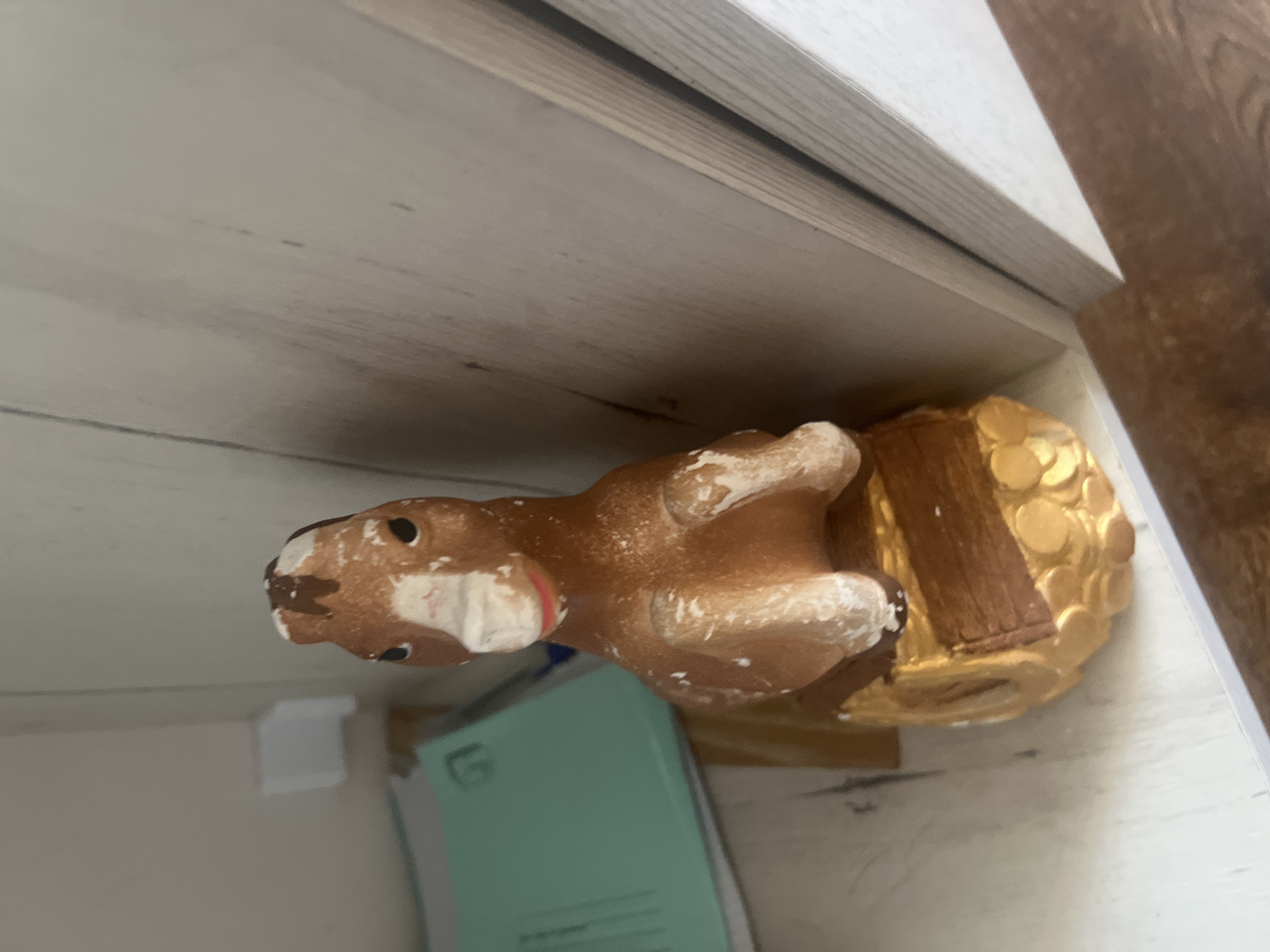 A small, brown and white ceramic horse figurine sits on a gold-colored base with wooden accents. The figurine has painted black eyes and a red mouth. It is positioned on a shelf next to a teal-colored envelope. The background consists of wooden shelving and a light-colored wall. The lighting appears to be natural, coming from the right side of the frame.