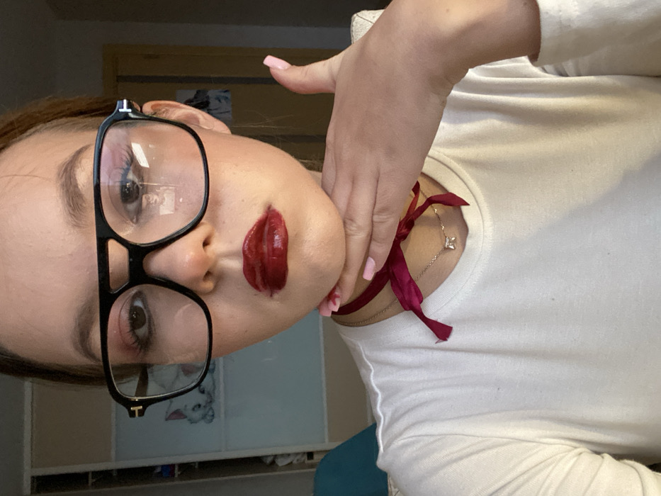 A close-up image depicts a young female indoors in Chaikovsky, Russia, captured from the forehead to the upper torso. She is wearing large black-framed aviator-style eyeglasses, dark red glossy lipstick, and eye makeup. A dark pink ribbon is tied around her neck, forming a bow beneath her chin. A thin silver chain necklace with a small, square clear gemstone pendant is visible below the ribbon. Her left hand is raised, with the palm resting against her chin and jawline, and her fingernails are painted light pink. She is dressed in a plain white long-sleeved shirt. The blurred background features a light-colored wardrobe or cabinet to the left, displaying a partial illustration of a white cat (Marie from "The Aristocats"). A reflection of a person's upper body is visible in the right lens of her eyeglasses.
