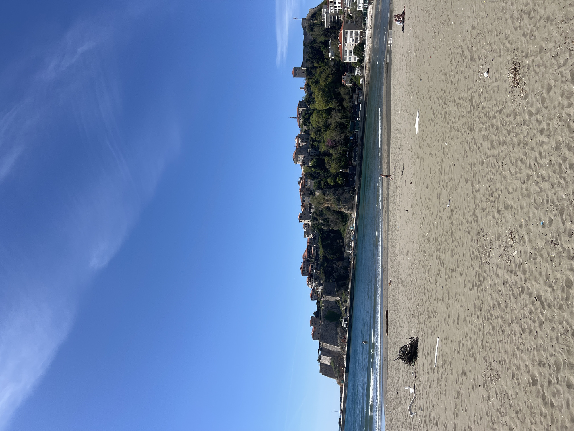 The image shows a coastline with a beach on the right and a hillside with buildings on the left. The sky is blue with some wispy clouds. The beach is sandy and has some debris scattered on it. The hillside is covered in greenery and has several buildings clustered along its slope. The water is calm and blue. The location is the city of Ulcinj - Ulqin, Montenegro.