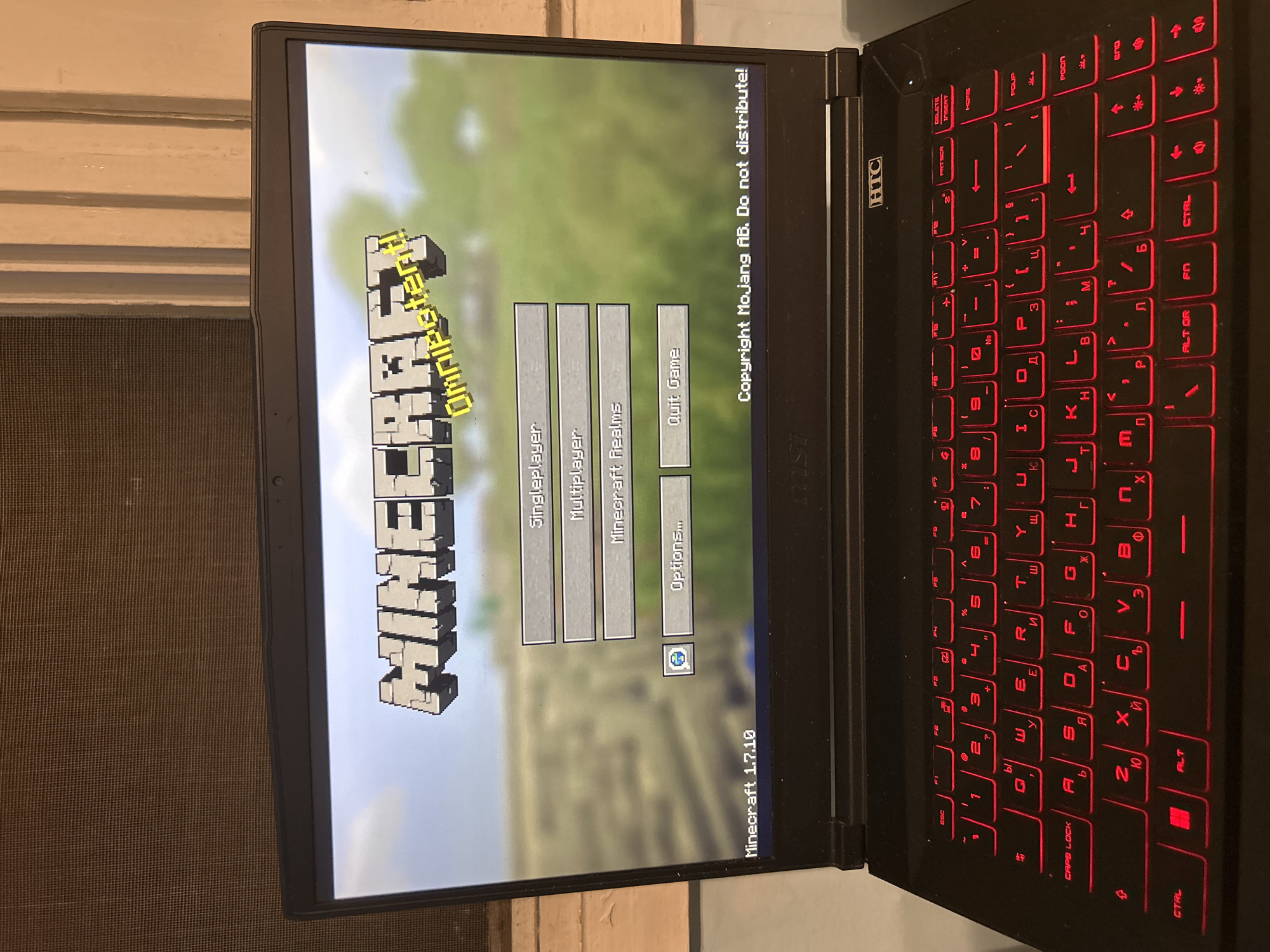 The image shows a laptop displaying the Minecraft main menu, with options such as "Singleplayer," "Multiplayer," "Minecraft Realms," "Options...," and "Quit Game." The Minecraft version is identified as 1.7.10. The laptop has a black chassis, and the keyboard features red backlighting with what appears to be Cyrillic script. The laptop's brand is "MSI". The surrounding environment is a window. The text on the screen includes the "Copyright Mojang AB. Do not distribute!" disclaimer. "Omnipotents!" is written above the "Minecraft" logo.