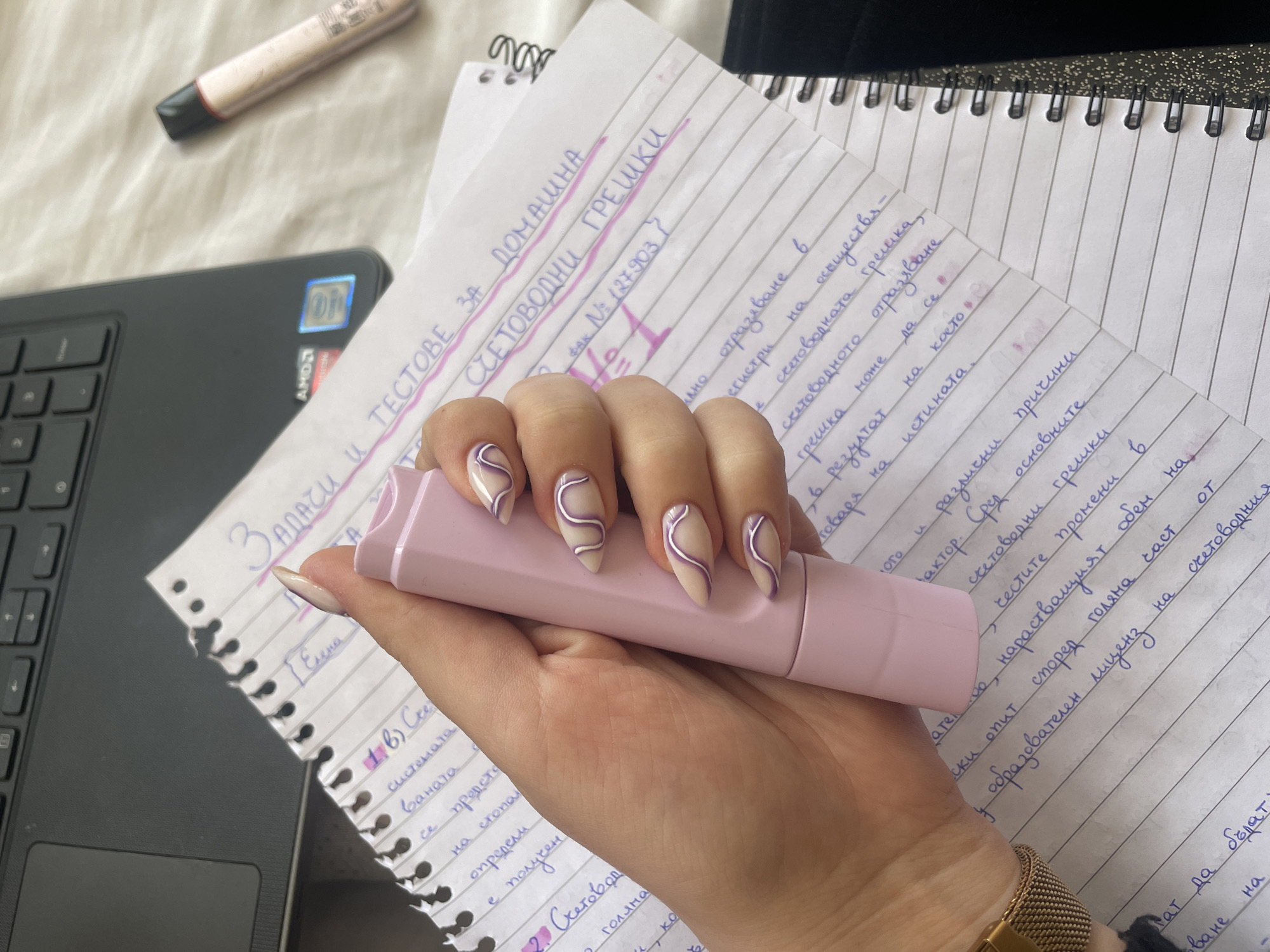 The image shows a close-up of a person's hand holding a pink object, likely a Kaweco Perkeo fountain pen or similar writing instrument. The person has long, almond-shaped nails with a light neutral base color and a curved purple line design. The hand is positioned over a notebook filled with handwritten notes in Bulgarian, possibly related to accounting or economics. The notebook is spiral-bound and rests on a black laptop. Also visible is a highlighter. The scene appears to be a study setting. The notes mention "домашни грешки" (domestic errors) and "счетоводни грешки" (accounting errors). It is impossible to tell that this is in the city of Varna, Bulgaria, since there is no location data.