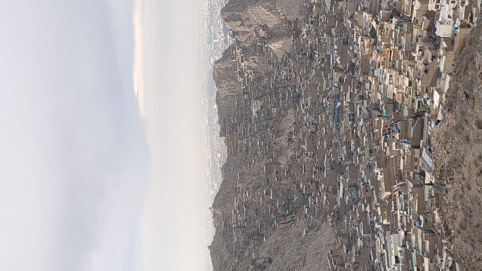 An aerial view captured from a high vantage point depicts a densely populated urban settlement in Quetta City Tehsil, Pakistan. The scene is dominated by a sprawling array of low-rise, flat-roofed buildings constructed directly onto and between arid, rocky mountains and hills. The structures are primarily light brown, beige, and grey, harmonizing with the natural tones of the terrain. Rooftops frequently feature blue cylindrical water tanks and various satellite dishes. The buildings exhibit an organic, irregular layout, conforming to the contours of the steep slopes. In the distant background, beyond the immediate cluster of buildings and mountains, the hazy expanse of the greater city of Quetta is visible under an overcast, light grey sky. No individuals, specific actions, or vehicles are discernible due to the elevated perspective and distance. The environment is characterized by a stark, arid landscape with minimal discernible vegetation.