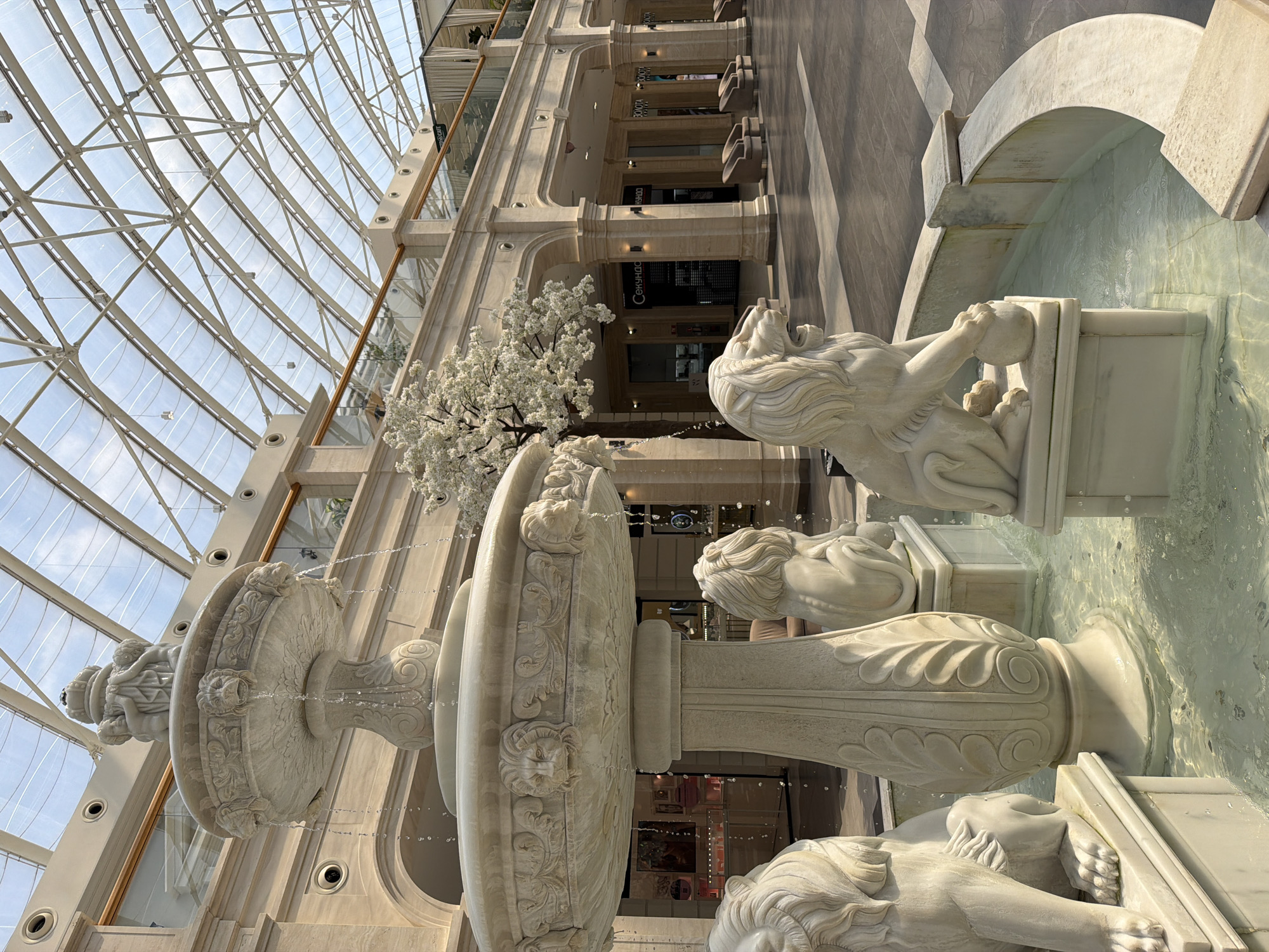 The image depicts the interior of a large, modern shopping mall, likely a public indoor space, with a grand, multi-tiered marble fountain as the central focal point. The fountain is intricately carved, featuring cherubic figures at the top and lion sculptures around its base, some holding spheres. Water cascades from various levels of the fountain into a pool. The mall's architecture is characterized by light-colored stone columns and arches, leading to a series of storefronts. Above, a transparent, arched ceiling allows natural light to illuminate the space, revealing the structural framework of the roof. A decorative, white-flowered tree stands to the right of the fountain, adding a touch of greenery. The floor is tiled in a geometric pattern. The overall atmosphere is one of opulence and public accessibility, characteristic of a high-end commercial or entertainment venue. There are no people visible in the scene.
The location is likely the Ocean Plaza mall in Kyiv, Ukraine, based on architectural similarities and the presence of similar decorative elements in publicly available images of the mall's interior, particularly the fountain.