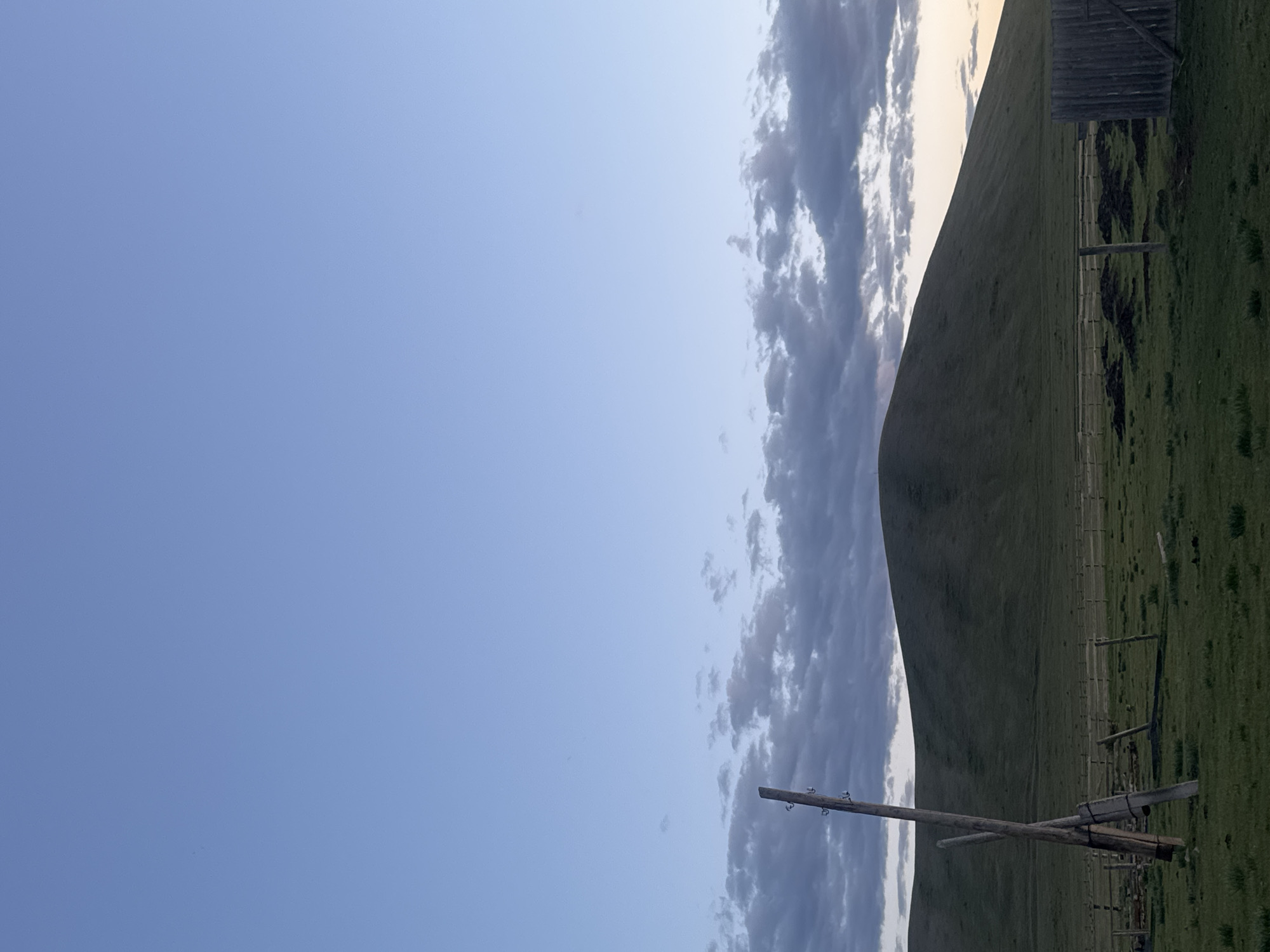 A landscape photograph, taken vertically, depicts a rural scene in Нижний Бургалтай, Russia. The sky occupies the left half of the image, transitioning from a clear blue to a band of streaky, gray clouds along the right edge. The right half shows a green, grassy hillside extending upward, dotted with sparse vegetation and sectioned off by a wooden fence line. A small, weathered wooden structure sits at the top of the hill. In the lower right corner, a fallen or dismantled utility pole lies on the ground, its wires absent. The light suggests either early morning or late evening, casting soft shadows across the terrain.