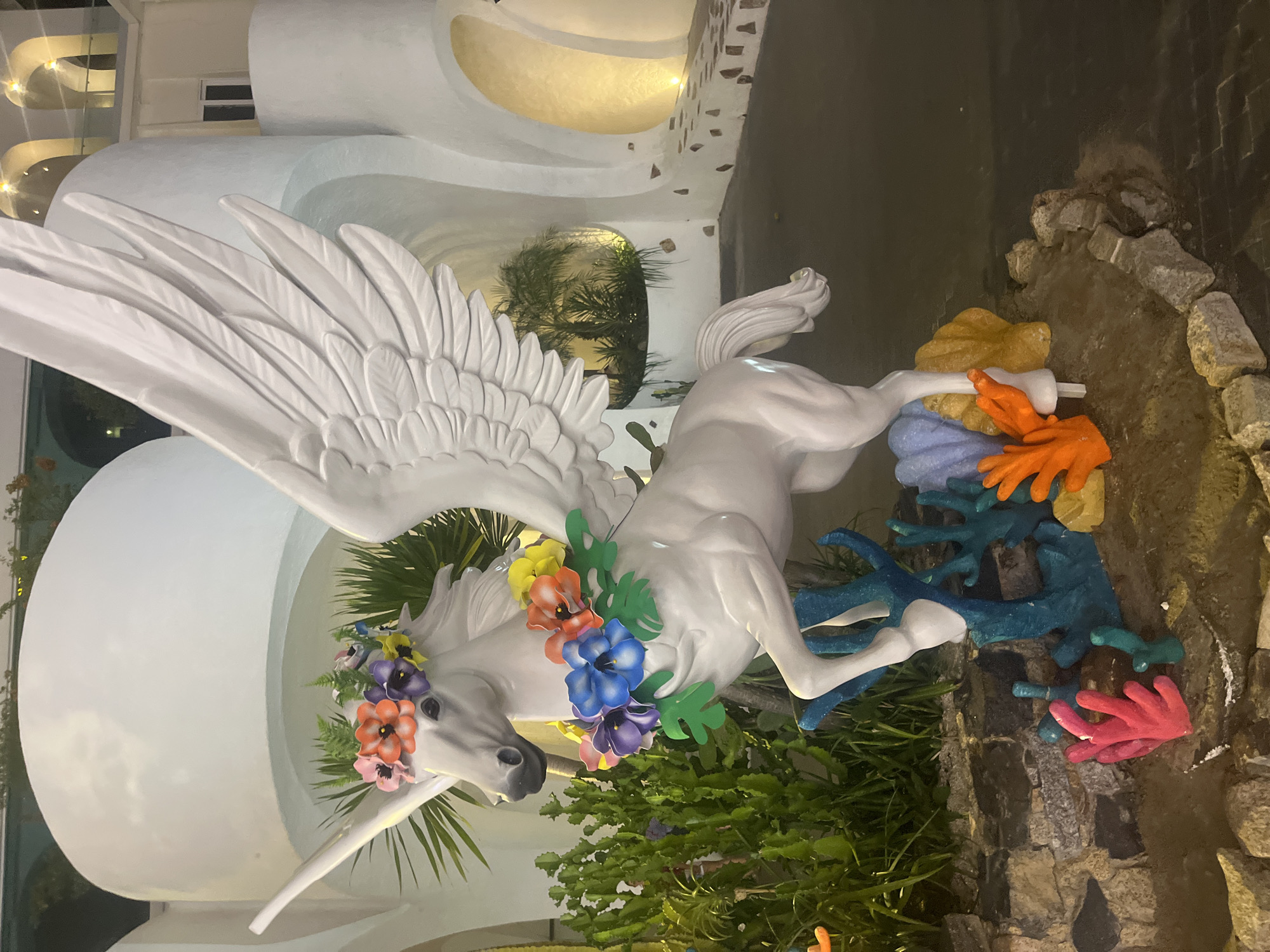 A white statue of a winged horse, possibly a Pegasus, is the central subject of this media. The statue is adorned with a colorful floral wreath around its neck and head, featuring flowers in shades of yellow, orange, pink, blue, and purple. Its wings are spread open, displaying detailed feather textures. The horse appears to be in mid-stride, with its front legs extended forward.
The statue is placed in an outdoor setting, possibly a garden or courtyard. Surrounding the base of the statue is a decorative element resembling coral, with vibrant colors like blue, orange, and pink. Some greenery, possibly palm fronds and other plants, is visible around and behind the statue. In the background, a white, modern building with curved architectural features and soft lighting can be seen. The overall ambiance suggests an evening or nighttime scene, given the artificial lighting present in the background and the slightly muted colors of the environment. The scene appears to be in Nha Trang Ward, Vietnam. There is no visible text in the image.