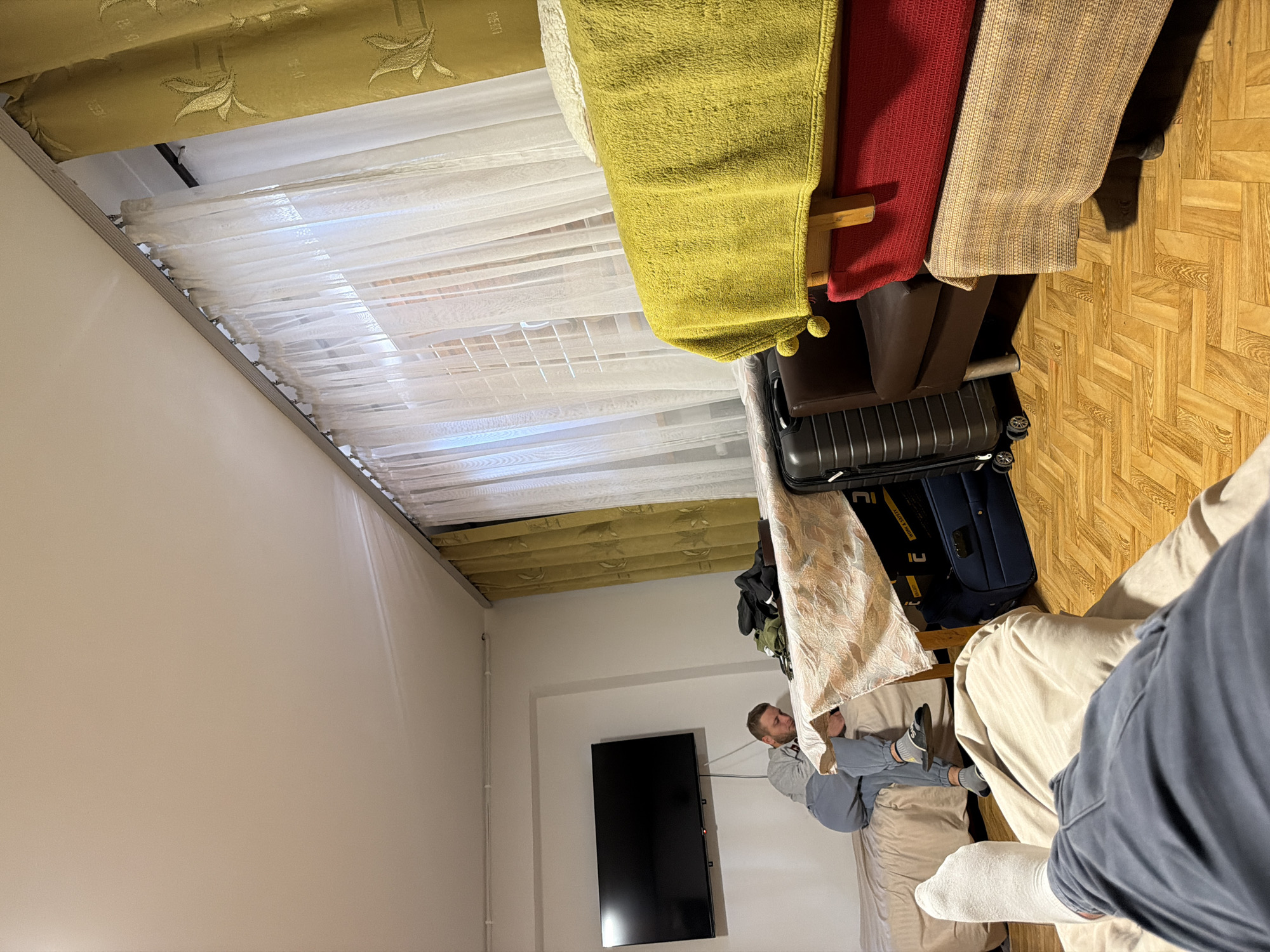 The image depicts an indoor scene, possibly a dormitory room or a shared living space, in Męcina, Poland. In the background, a man in a grey hoodie and blue pants is sitting on a bed, looking down at his phone. A black television is mounted on the wall above him. In the foreground, another person's legs, clad in blue pants and white socks, are visible, suggesting they are lying down.
The room features wooden parquet flooring. A table with a patterned tablecloth is centrally placed, covered with various items including dark clothing and a green backpack. Beside the table, a brown armchair is visible, with several suitcases stacked next to it. The furniture appears functional and somewhat utilitarian. The room is illuminated by ambient light, with light filtering through sheer white curtains and heavier yellow curtains hanging near a window. The overall impression is of a lived-in, temporary living arrangement.