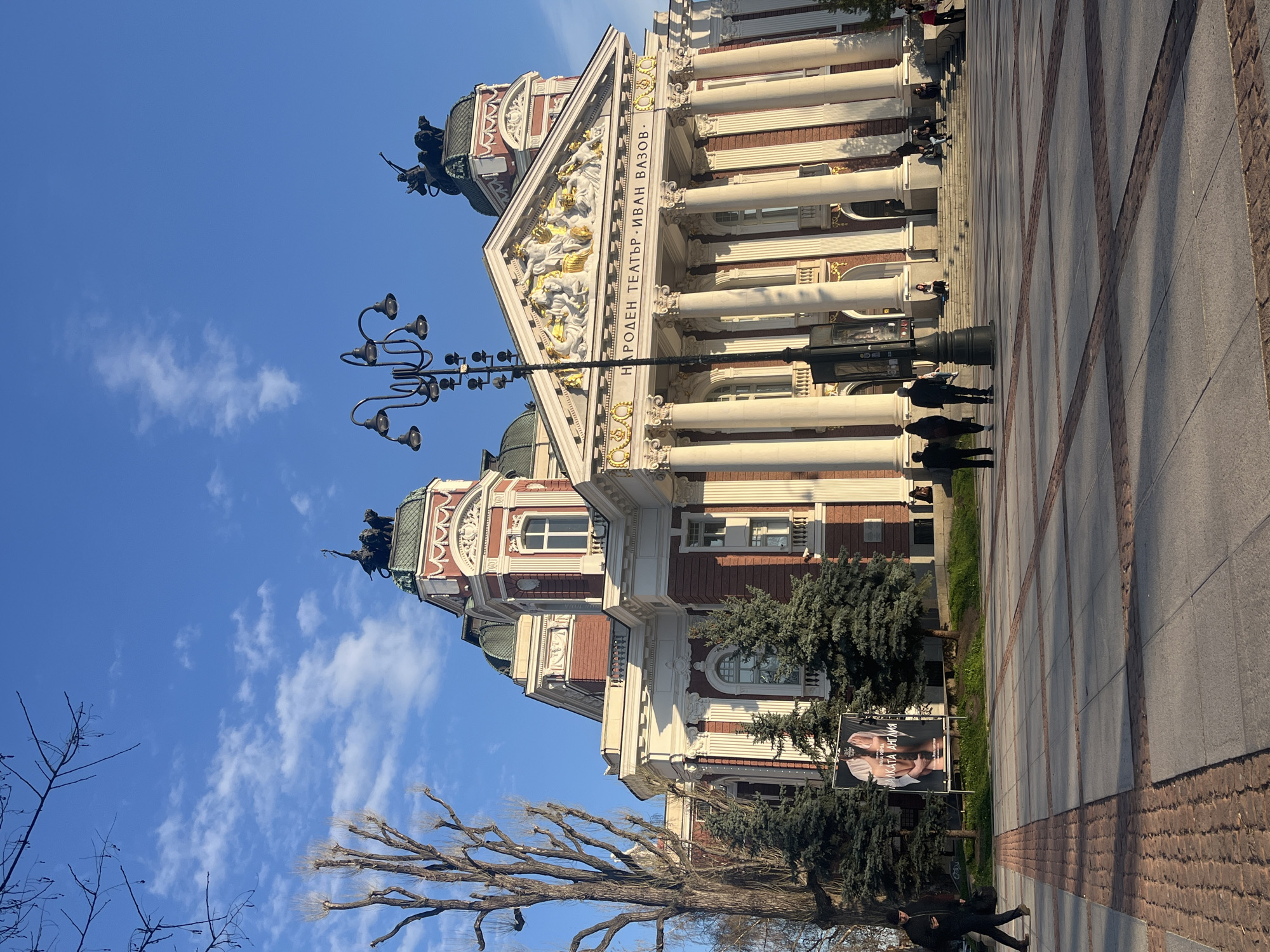 The image captures the Ivan Vazov National Theatre in Sofia, Bulgaria, taken from a ground-level perspective, depicting the building's facade against a blue sky scattered with clouds. The theatre features a classical architectural style with tall white columns, ornate gold detailing, and a pediment adorned with sculptural figures. In front of the building, pedestrians are visible on a paved plaza. A vintage-style lamppost stands near the building, adding to the scene's historical ambiance. The foreground includes bare tree branches, suggesting it might be late autumn or early spring. A poster is visible in front of the building. The overall atmosphere is sunny and clear, highlighting the architectural details of the theatre and the surrounding urban environment.