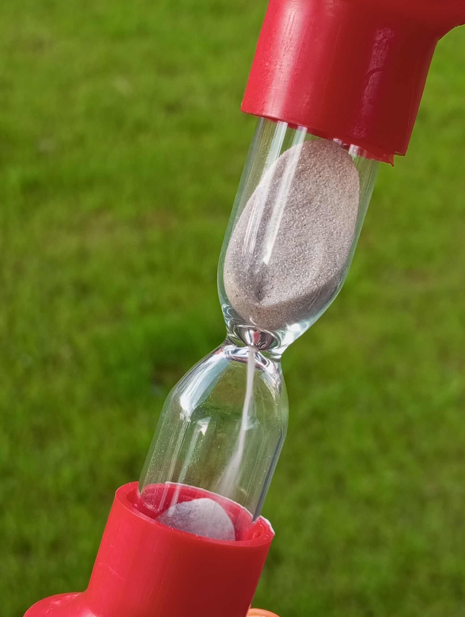 A close-up shot features a small hourglass set against a vibrant, out-of-focus green grassy background, suggesting an outdoor setting in Pervomaysky, Russia. The hourglass consists of clear glass chambers encased by bright red plastic caps at its top and bottom. Light grey sand is actively flowing from the upper chamber to the lower, indicating the timer is in operation. A substantial amount of sand remains in the upper bulb, while a smaller, growing pile is visible in the bottom. The hourglass is positioned diagonally across the frame under bright natural light, implying daytime. No individuals are present, and no text is visible on the hourglass or in the background.