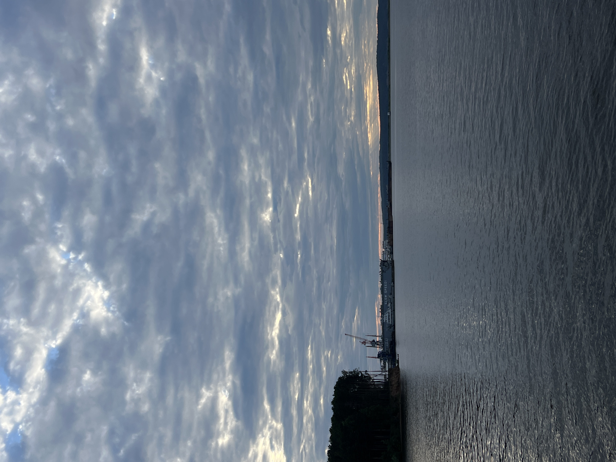 The image depicts a wide, calm body of water, likely the Kama River, with a shoreline on the left featuring a dense forest of dark green trees. Adjacent to the forest, along the water's edge, is a shipyard or industrial area with several large structures. These include a prominent red and white crane, a large grey vessel or building labeled "SLOW SPEED," and other docked boats and industrial equipment. The water's surface is slightly rippled, reflecting the overcast sky.
The sky is filled with a textured pattern of grey and white clouds, with patches of sunlight breaking through, suggesting either early morning or late afternoon. The horizon in the distance shows a low, dark landmass, possibly hills or a distant shoreline. The overall impression is a serene yet industrial waterscape, characteristic of the port areas found in Chaikovsky, Russia, a city known for its hydroelectric power station and industrial enterprises on the Kama River. No people are visible in the frame.
