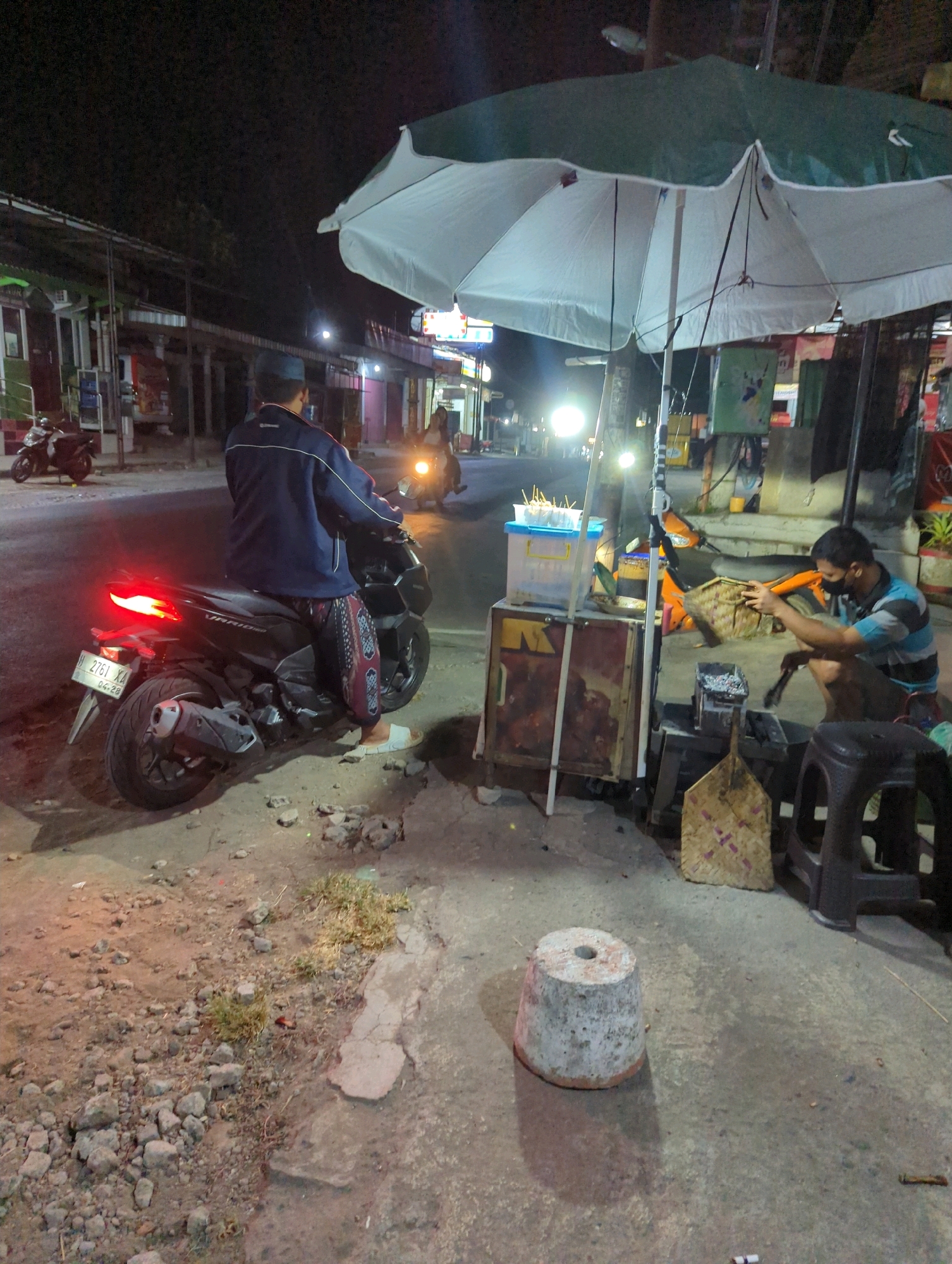 In Kacangan, Indonesia, at night, a street scene unfolds under a large white and green umbrella. A person wearing a blue jacket and patterned pants sits on a black motorcycle, its taillight illuminated. The motorcycle has a license plate reading "2761 XA 04-28". To the right, a man with a black face mask, wearing a blue and white striped shirt, crouches by a small charcoal grill. He is holding a woven basket and a tool, appearing to be preparing food. In front of him is a food cart with a blue container holding skewers and an orange motorcycle parked behind him. The ground is a mix of dirt, gravel, and cracked concrete. In the background, other motorcycles are visible, as well as the facades of shops illuminated by streetlights. The overall atmosphere suggests a bustling night market or roadside food stall area.