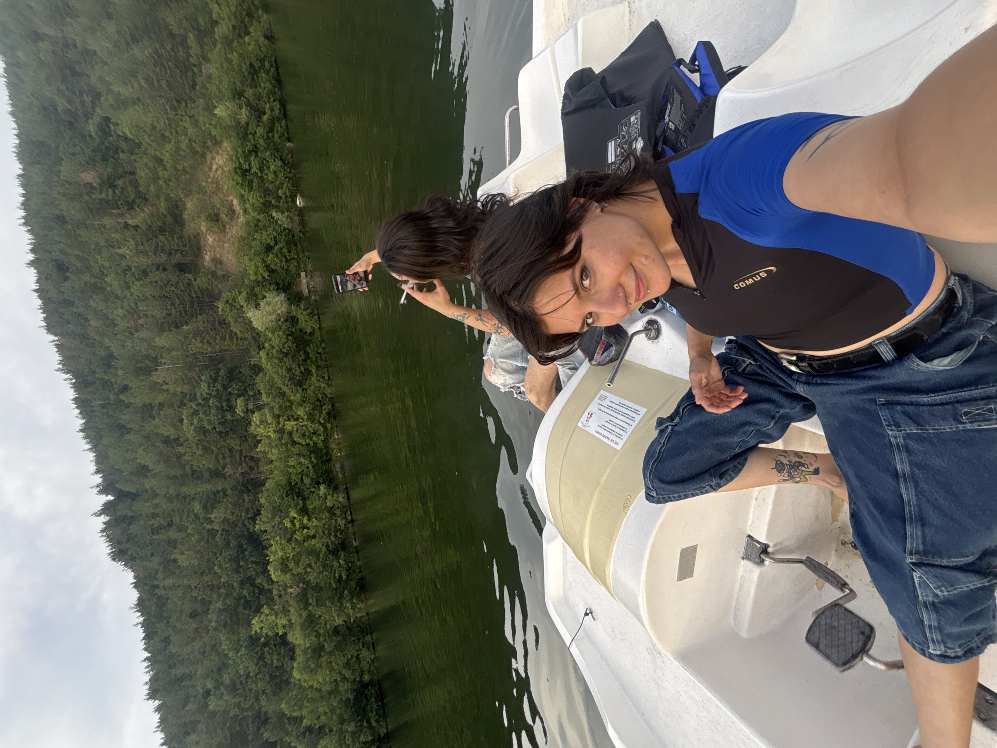 Two women are in a pedal boat on Lake Pancharevo, Bulgaria. The woman in the foreground is taking a selfie, smiling at the camera. She has dark hair, a nose ring, and is wearing a black and blue cropped shirt and blue jeans. She has a tattoo on her left leg. The woman in the background is looking at her phone and smoking. She has dark hair and is wearing ripped blue jeans. The pedal boat is white with a beige seat. The lake is surrounded by a dense forest of green trees. The sky is overcast.