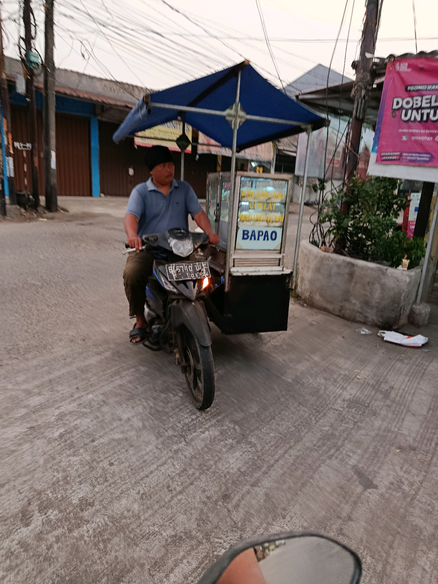 A man wearing a blue polo shirt and a black cap is riding a motorcycle with a food cart attached. The food cart has a blue canopy and displays the word "BAPAO" in large letters. There are also some other items visible in the cart. The motorcycle is dark gray and has a license plate that reads "B 5710 UIE". The man is looking forward, and his hands are on the handlebars. The motorcycle is on a paved street, and there are buildings and power lines in the background. A pink banner with Indonesian text is visible on the right side of the image. The scene appears to be a street in the city of Bekasi, Indonesia, likely in an urban or suburban area.
