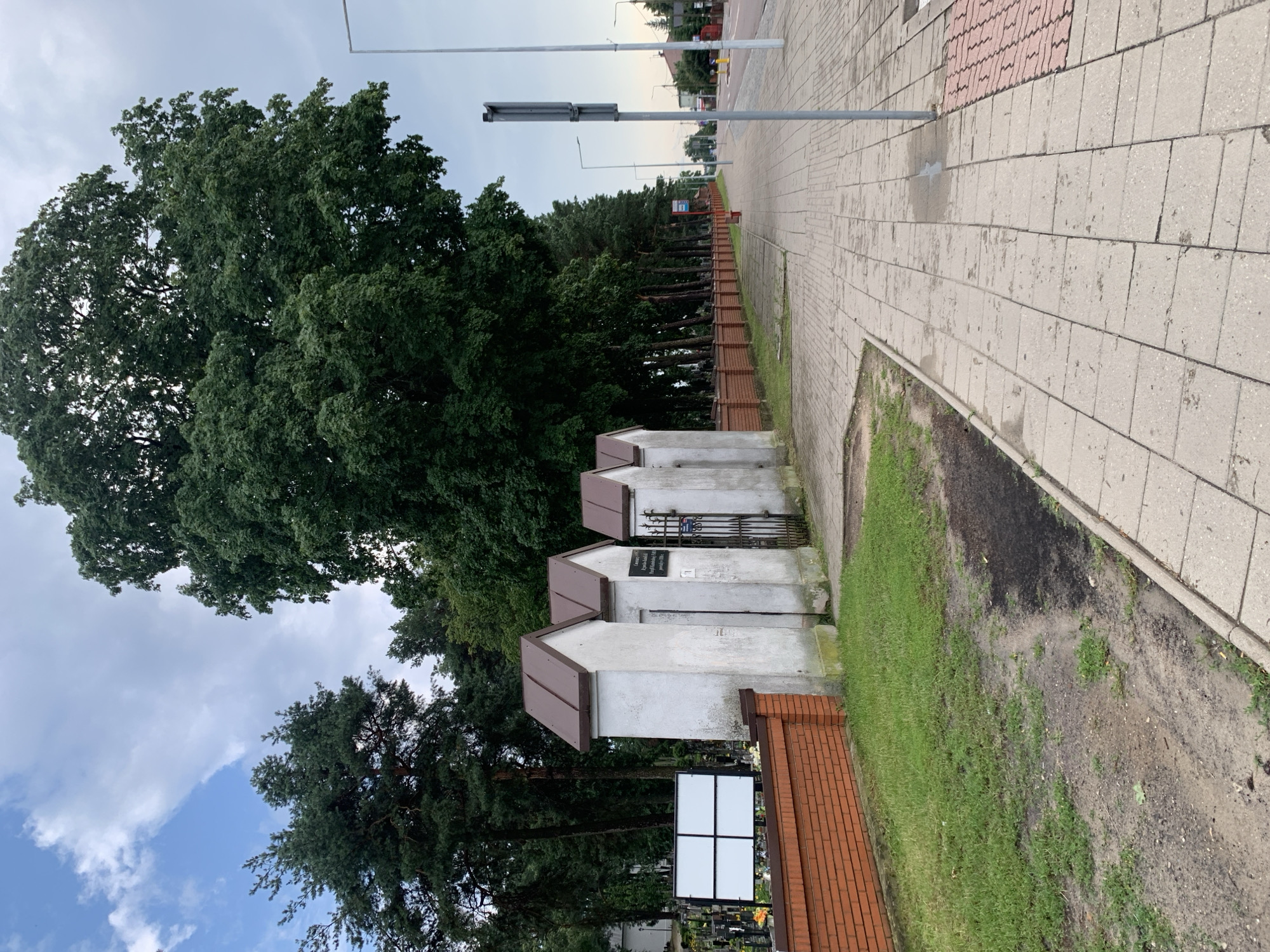 The image shows a street scene in Białystok, Poland, with a sidewalk and a cemetery on the left. The cemetery features a brick wall and several small, white structures with brown roofs. A large, leafy tree dominates the background, along with a line of evergreen trees. The sidewalk is paved with concrete blocks, and there are streetlights and a road visible further down. The sky is partly cloudy. There are no people visible in the image.