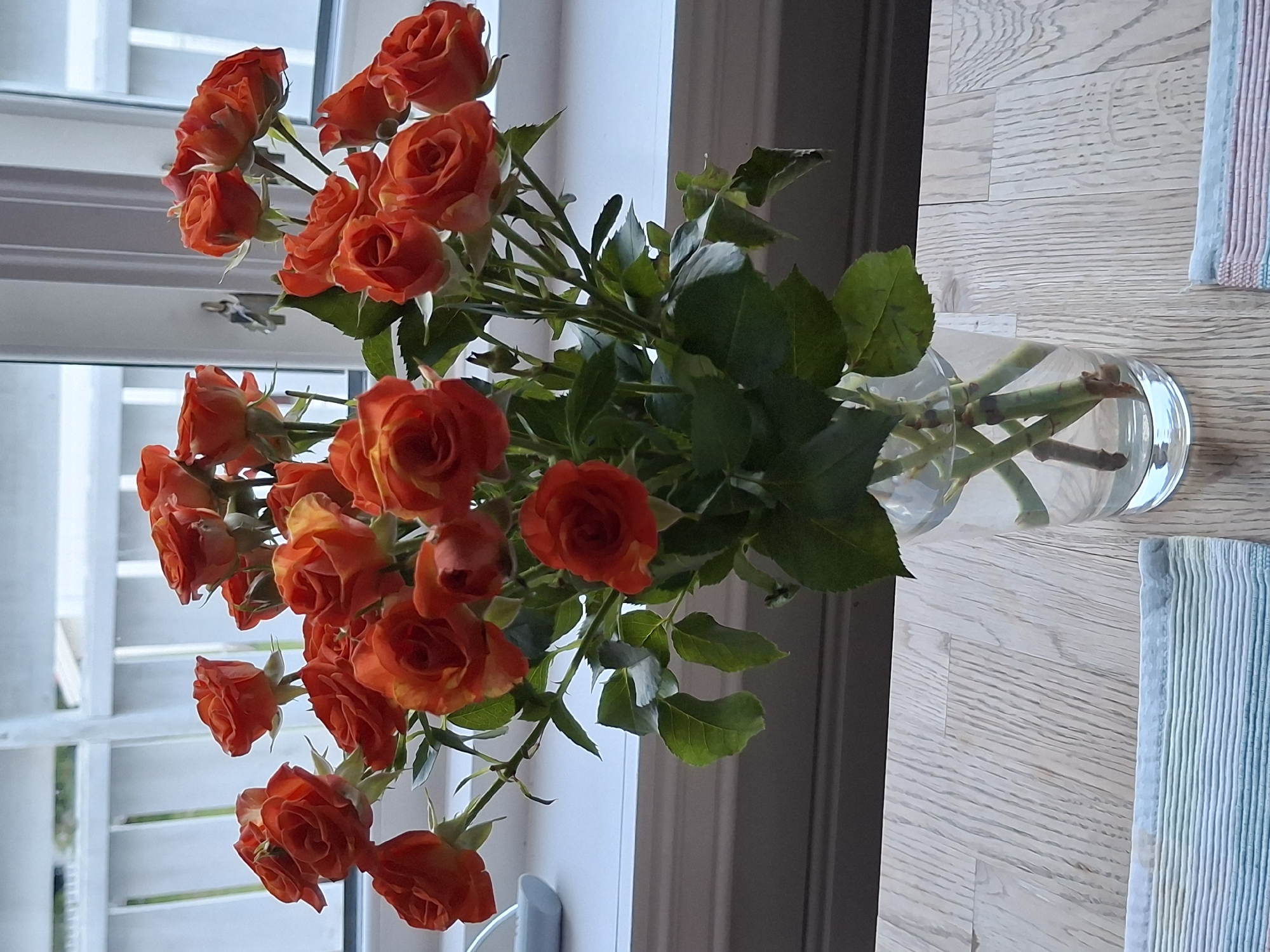 A bouquet of orange roses with green leaves is displayed in a clear glass vase filled with water. The vase sits on a light-colored wooden surface. In the background, there is a white-paneled window with a view of a white fence or railing. The overall scene is well-lit, suggesting it is daytime. The location in the city of Åsgårdstrand, Norway, is an indoor setting, likely a home or a business, near a window that looks out onto a residential or garden area.