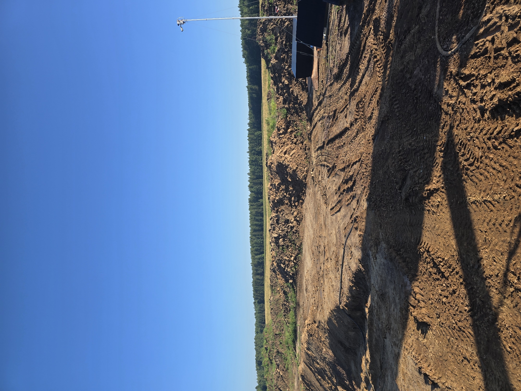 This image depicts an outdoor construction site under a clear blue sky. The foreground is dominated by disturbed earth and tire tracks, indicating recent activity by heavy machinery. In the mid-ground, there are piles of dirt and some green vegetation. Beyond that, a line of trees forms the horizon. To the right of the image, a tall pole with a camera and some dark, rectangular structures are visible, suggesting temporary site security or monitoring equipment. The location is identified as Piecnik, Poland. There are no people visible in the image.