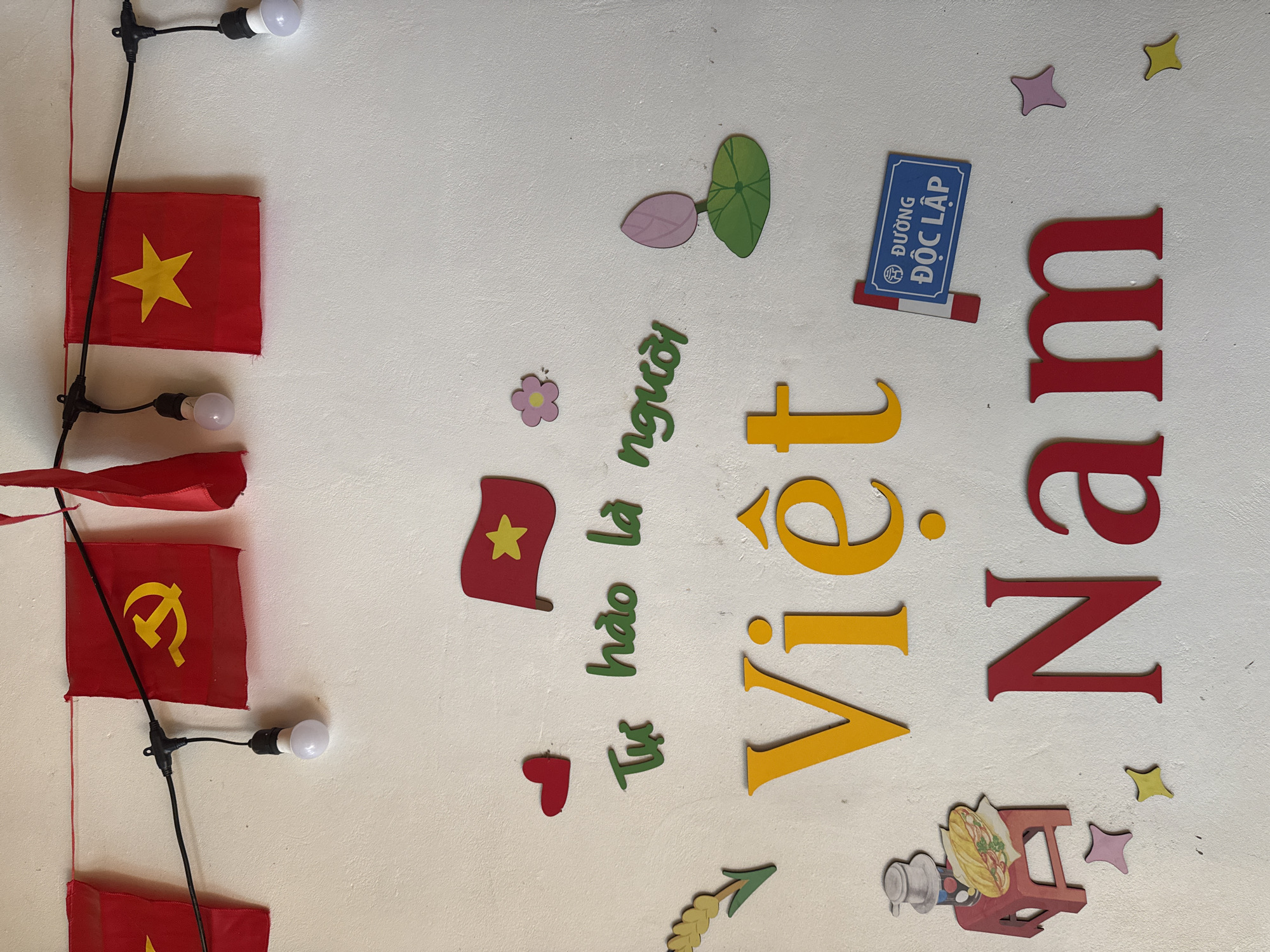 The image shows a white wall adorned with Vietnamese decorations. A string of lights with red Vietnamese flags is strung along the top left of the frame. Decorative elements on the wall read "Việt Nam" in large, colorful letters, and the phrase "Tự hào là người" in green. There is a lotus flower symbol and a blue sign that says "Đường Độc Lập". There is also a small image of a sandwich and a drink. The scene is set in Púng Luông, Vietnam.