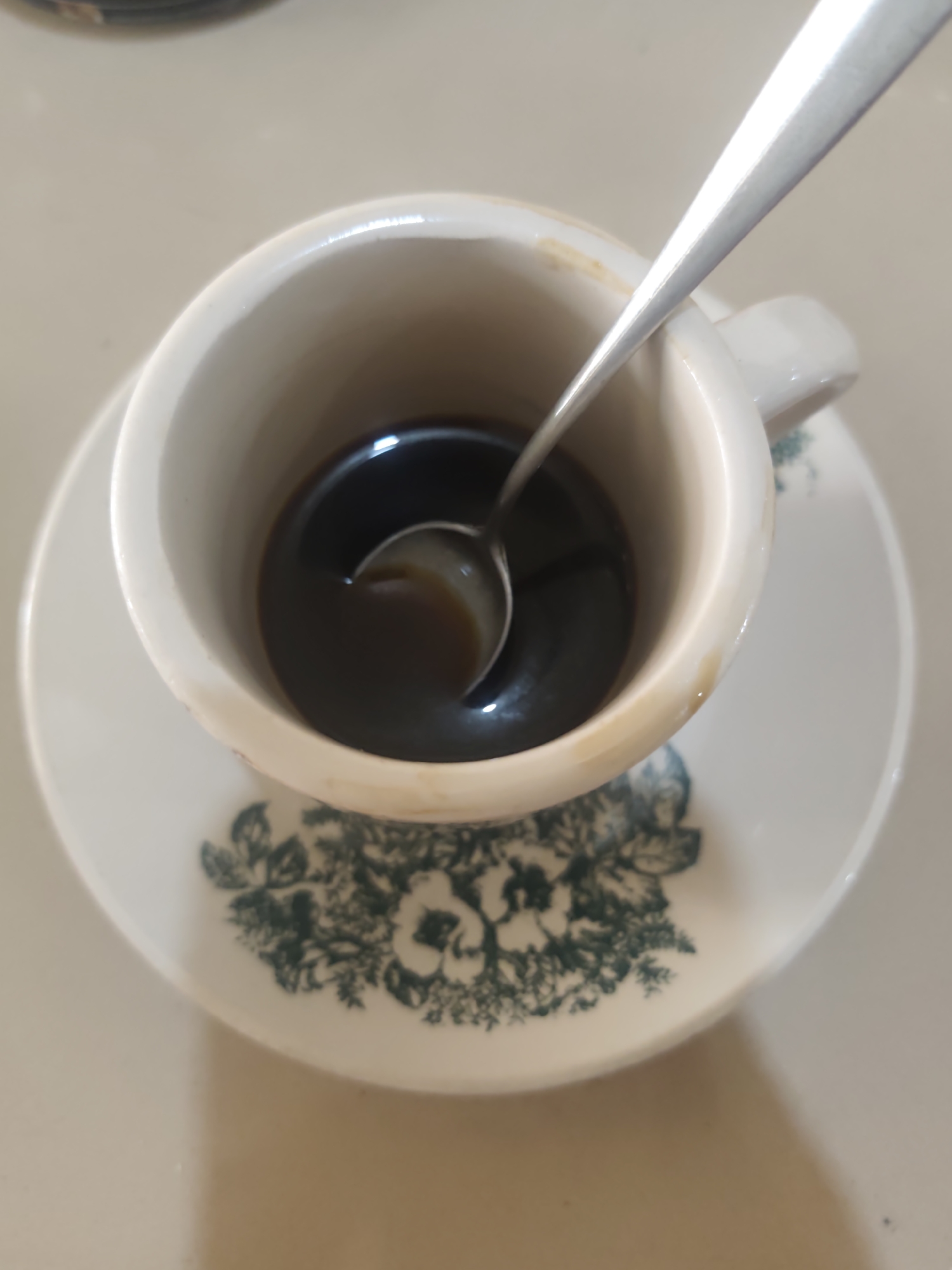 A close-up overhead shot shows a white ceramic coffee cup with a metallic spoon resting inside. The coffee within the cup is dark brown. The cup sits on a matching white saucer adorned with a dark green floral pattern. The background is a neutral, light-colored surface. There are no people or animals visible in the frame. The scene appears to be a simple, domestic setting, possibly a kitchen or dining area in a home in Pematang Reba, Indonesia, though no specific landmarks are identifiable.