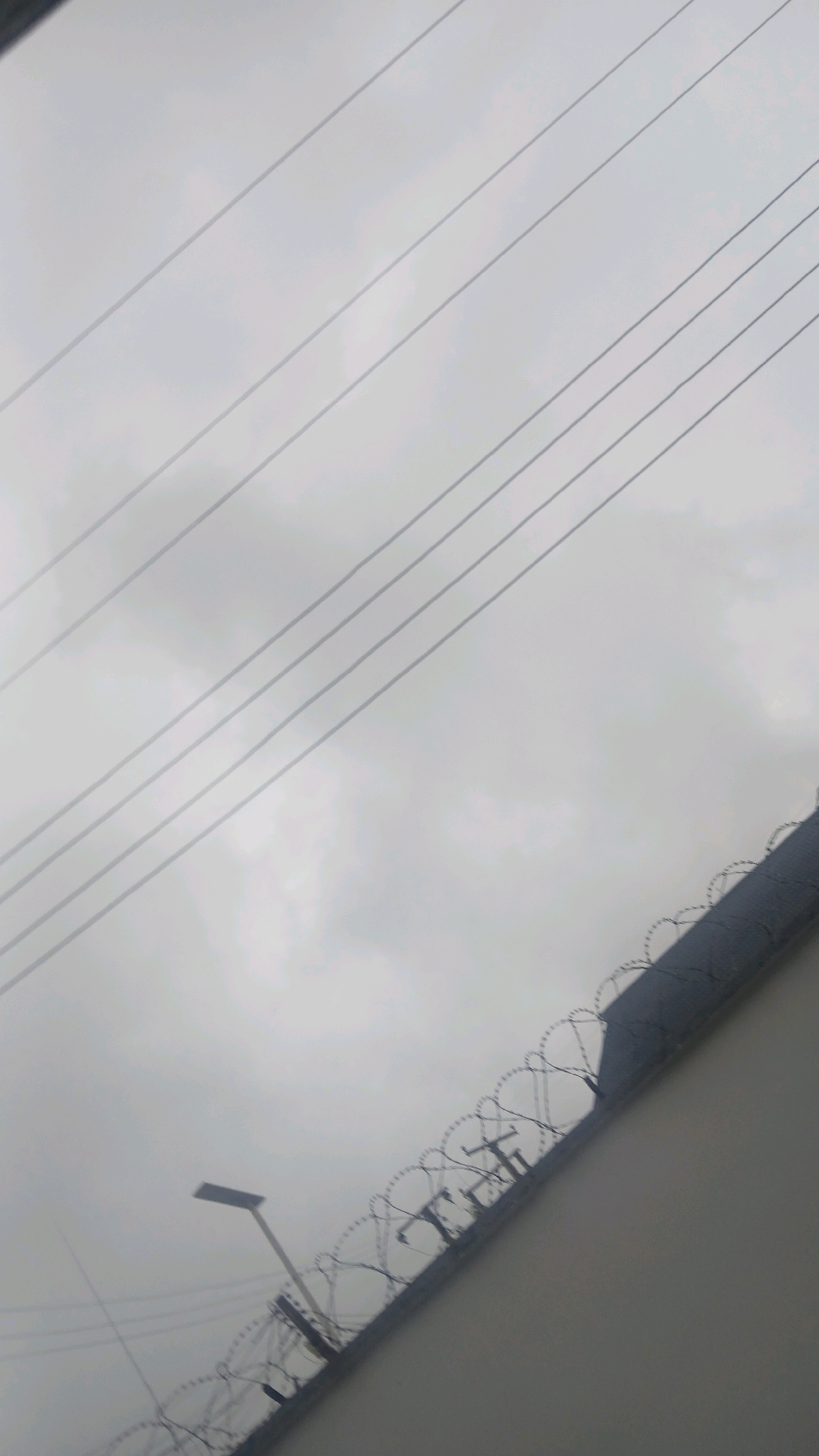 The image depicts an overcast sky above a high concrete wall topped with barbed wire fencing. Several electrical wires run diagonally across the upper portion of the frame. A street lamp is visible to the left, partially obscured by the barbed wire. The scene appears to be an outdoor environment, possibly a perimeter fence in an urban or semi-urban setting. Based on the available information, the location is Gura Top, Nigeria. There are no people or discernible activities in the image.