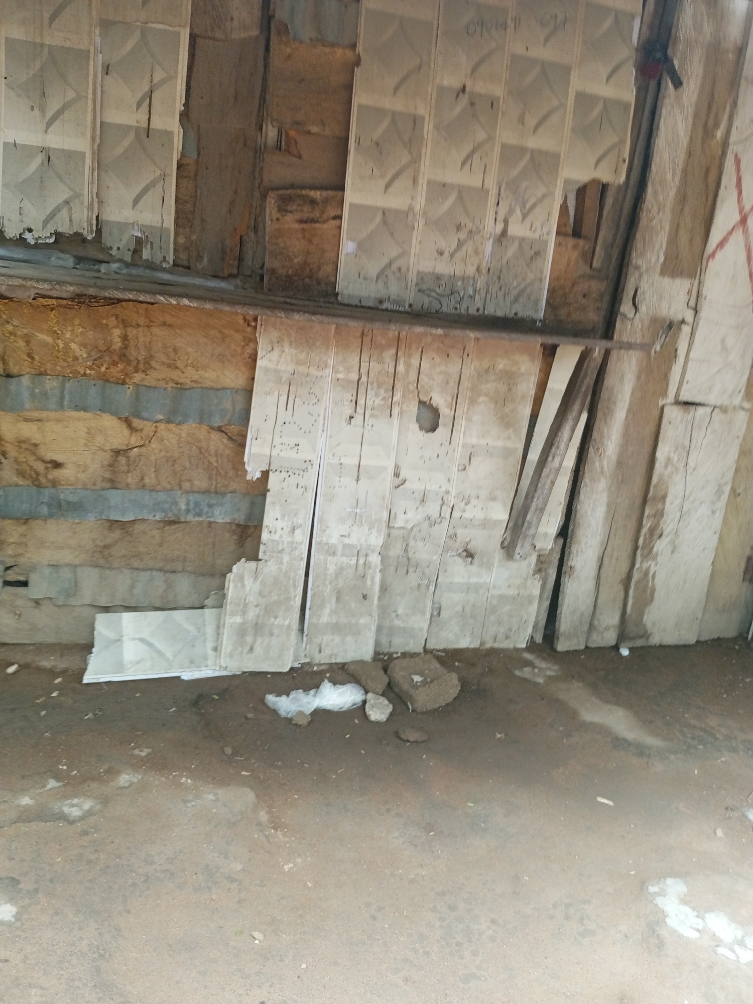 The image depicts an outdoor scene in Jalingo, Nigeria, likely a storage area or workshop. The primary subjects are building materials, specifically several stacks of what appear to be large, light-colored tiles or panels, some showing wear and tear. These are leaning against a rough wooden structure that forms the background. The wooden structure itself is composed of various planks and possibly corrugated metal strips, suggesting a rustic or makeshift construction.
In the foreground, the ground is a dirt or gravel surface, appearing somewhat uneven and damp in places, with scattered debris such as small stones and white plastic fragments. One stack of tiles is laid flat on the ground in the lower left corner.
There are no people visible in the image. The activity taking place, if any, is not directly depicted, but the presence of building materials suggests a potential construction, renovation, or storage scenario.
Notable details include the textured surface of the tiles, which have a diamond or geometric pattern. Some of the tiles show signs of age, with chipped edges and discoloration. The wooden structure behind the tiles is weathered and shows grain and cracks. The lighting suggests it could be daytime, but there are no strong shadows to indicate a specific time. The overall impression is one of a utilitarian and somewhat neglected outdoor space.
No discernible text is visible in the image, other than faint markings on some of the tiles that are illegible.