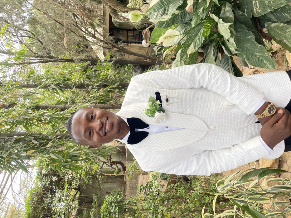 An outdoor, eye-level, mid-shot image captures a male subject, of apparent African descent, centered and smiling directly at the viewer. He presents with a short, dark haircut and a clean-shaven face. The subject is formally dressed in a white, textured (possibly pique or waffle weave) tuxedo jacket featuring a single button closure and shawl lapels, worn over a white dress shirt. A black velvet bow tie is positioned at his neck. A white boutonnière with green foliage is pinned to his left lapel, and a black pocket square, secured by a decorative pin with a dark stone and two silver chains, is visible in his left breast pocket. A gold-toned wristwatch is worn on his left wrist, and his hands are clasped together at waist level. The background consists of dense, green vegetation, including tall, slender trees with drooping leaves to the left and a large plant with variegated green and white leaves and white flowers to the right. A rustic structure, possibly a hut or gazebo with a natural material roof, is partially visible in the upper right background. The scene is illuminated by natural daylight. While the location is specified as Jos, Nigeria, no specific landmarks are discernible within the image to pinpoint an exact site or address in the city.