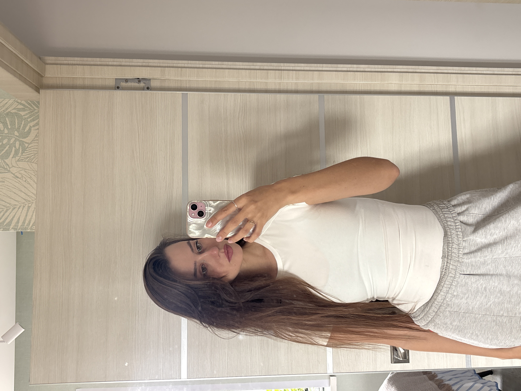 A woman is taking a selfie in front of a mirror. She is wearing a white T-shirt and grey sweatpants. The background includes a beige wooden door with metal accents and a wall with leaf-patterned wallpaper. The photo was taken indoors. There is no indication of the location being Krasnoyarsk, Russia.