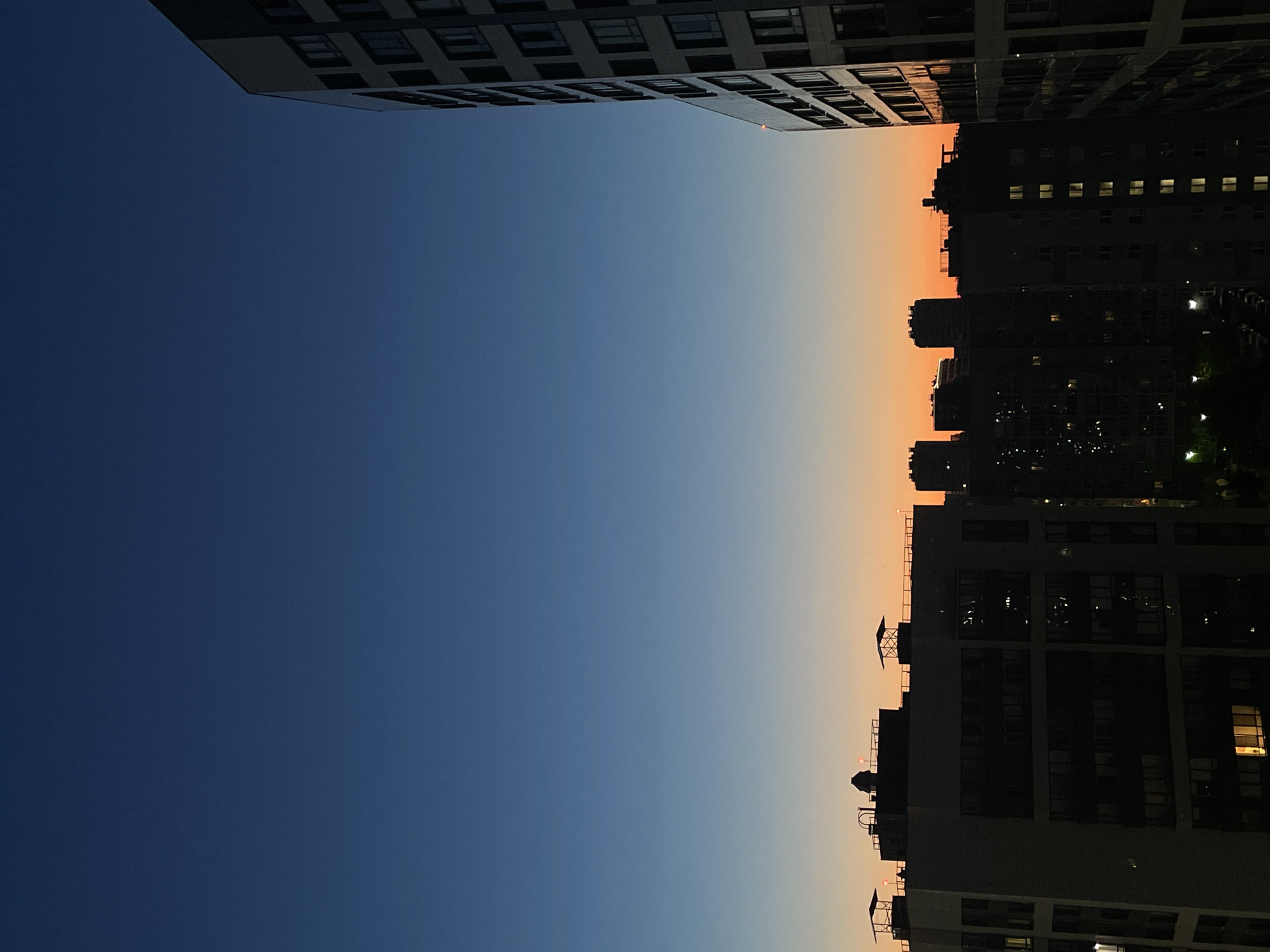 The image depicts a city skyline at dusk in Sinyavino, Russia. Silhouetted against a gradient sky transitioning from deep blue to a soft orange hue are several multi-story buildings. The structures are dark and feature visible windows, some of which are illuminated, suggesting interior lighting. On the rooftops of some buildings, distinct architectural elements such as vents and what appear to be small roof structures are discernible. A tall, modern building with a repetitive window pattern is prominent on the right side of the frame, its facade catching the last light of the day. The overall scene is one of urban development under a fading twilight sky, with no people or active events visible.