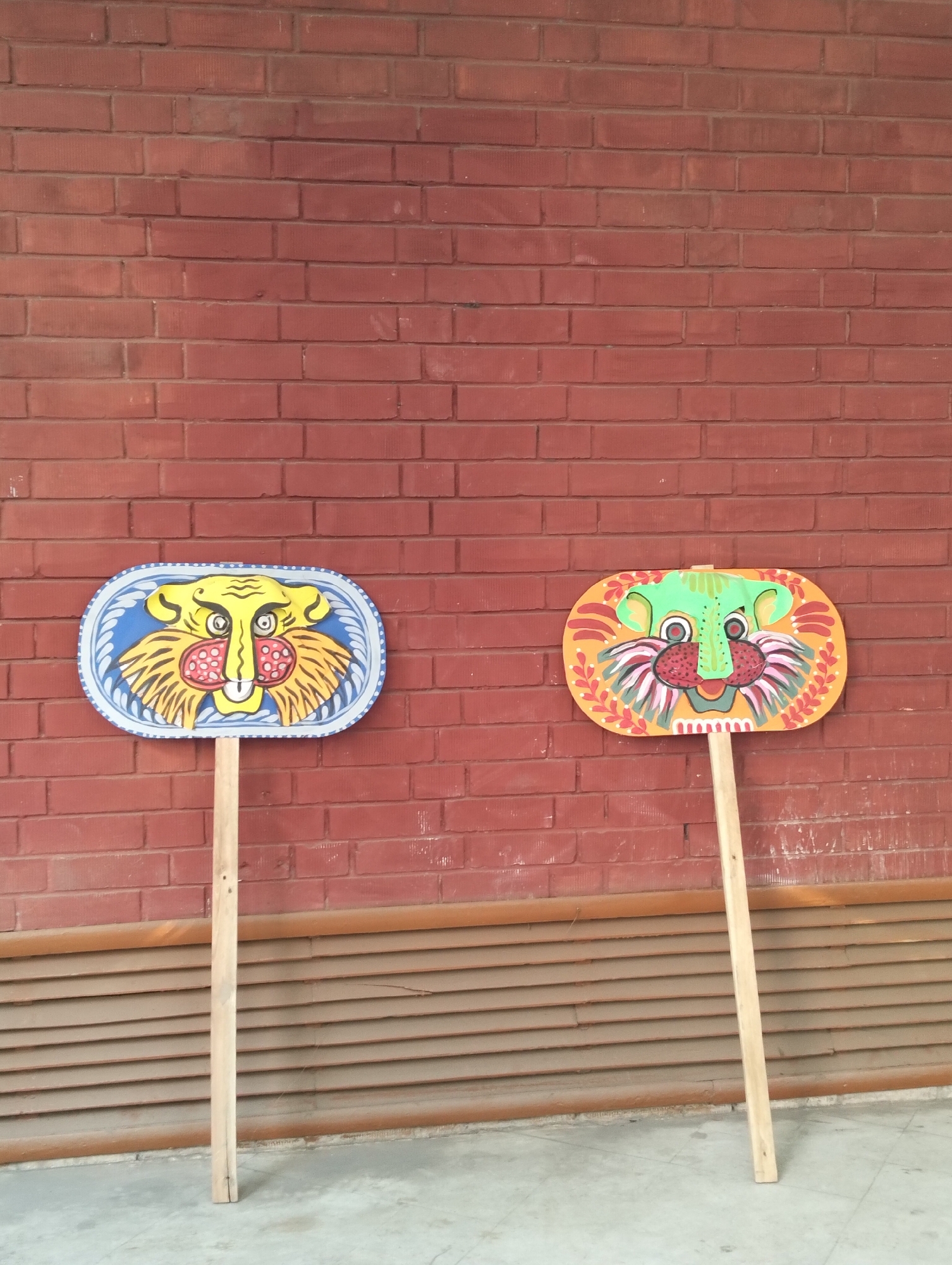 The image depicts two painted tiger masks attached to wooden sticks, standing against a red brick wall and a horizontal slatted partition. The left mask has a yellow tiger face with a blue border, while the right mask features a green tiger face with an orange border. The masks appear to be standing on a concrete surface. The location appears to be outdoor, potentially a street in the city of Unknown, Unknown.