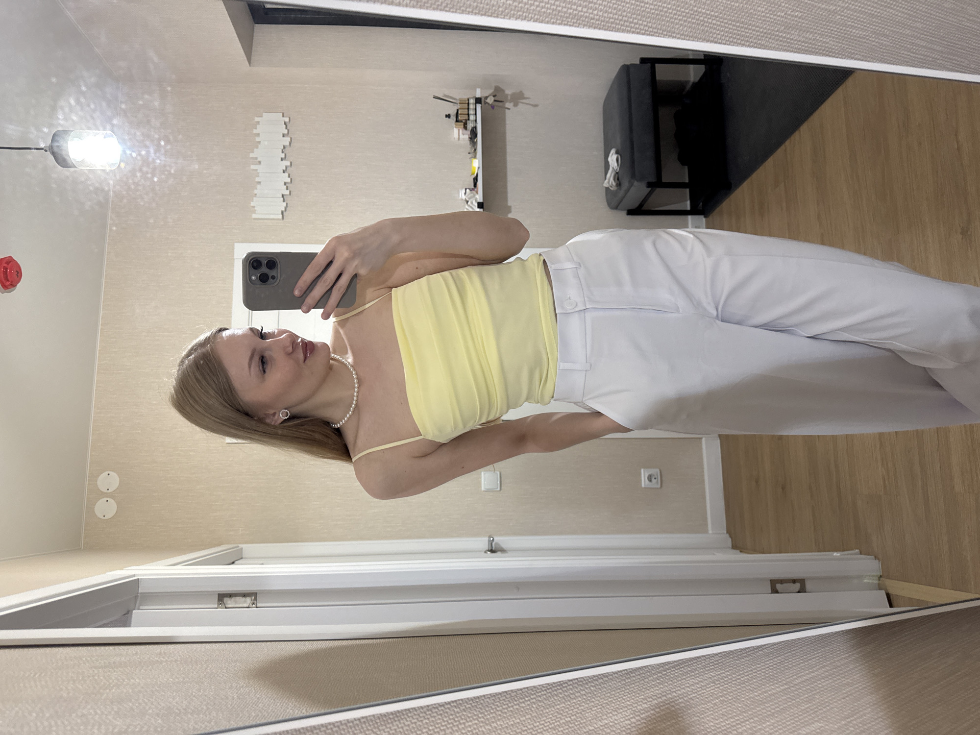 A young woman with blonde hair takes a mirror selfie in what appears to be an indoor setting. She is wearing a light yellow camisole with spaghetti straps and high-waisted white trousers. A pearl necklace adorns her neck, and she has a small stud earring in her visible ear. She holds a dark-colored smartphone in her right hand, angled to capture her reflection. The background reveals a neutral-toned wall, a wooden floor, and part of a white door frame. To the left, a cylindrical light fixture with a bright bulb is visible, casting a glare and creating a starry effect on the wall. Further back, a shelf with various items and a dark upholstered bench are present. The image suggests a casual moment of self-presentation, possibly at home. The lighting indicates it could be daytime, but the lack of natural light sources and presence of artificial illumination makes it difficult to determine the exact time. There is no visible text in the image.
