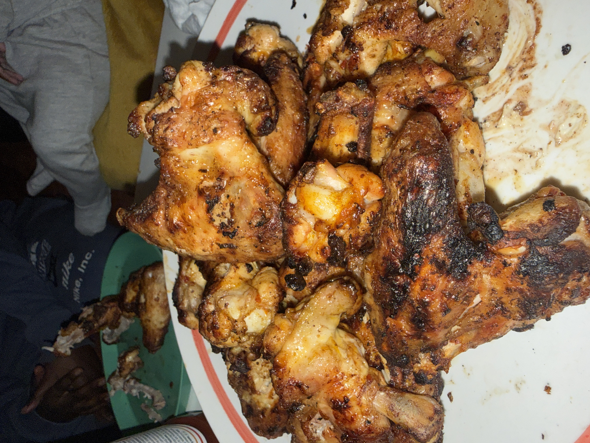 A close-up shows a platter piled high with cooked chicken wings. Some wings appear golden brown and crispy, while others are charred. A side plate holds remnants of consumed wings, consisting of bones. A person wearing a grey Nike t-shirt is partially visible. The photo was taken indoors. No geolocation information is provided, so the location cannot be verified.
